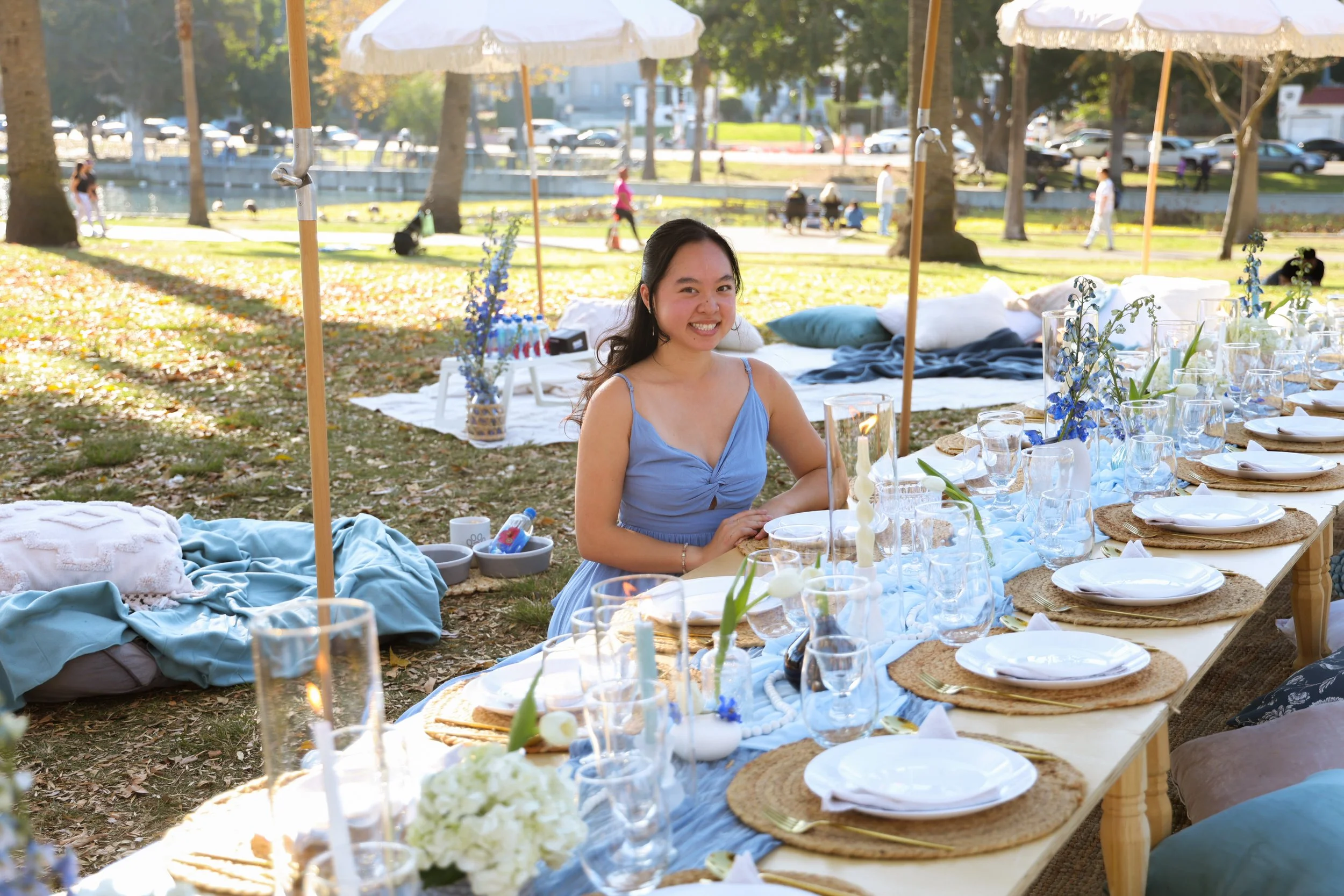Young woman sitting at an outdoor dining table with a smile, set for a meal with plates, glasses, and floral decorations, shade umbrellas in the background, in a park setting during daytime.