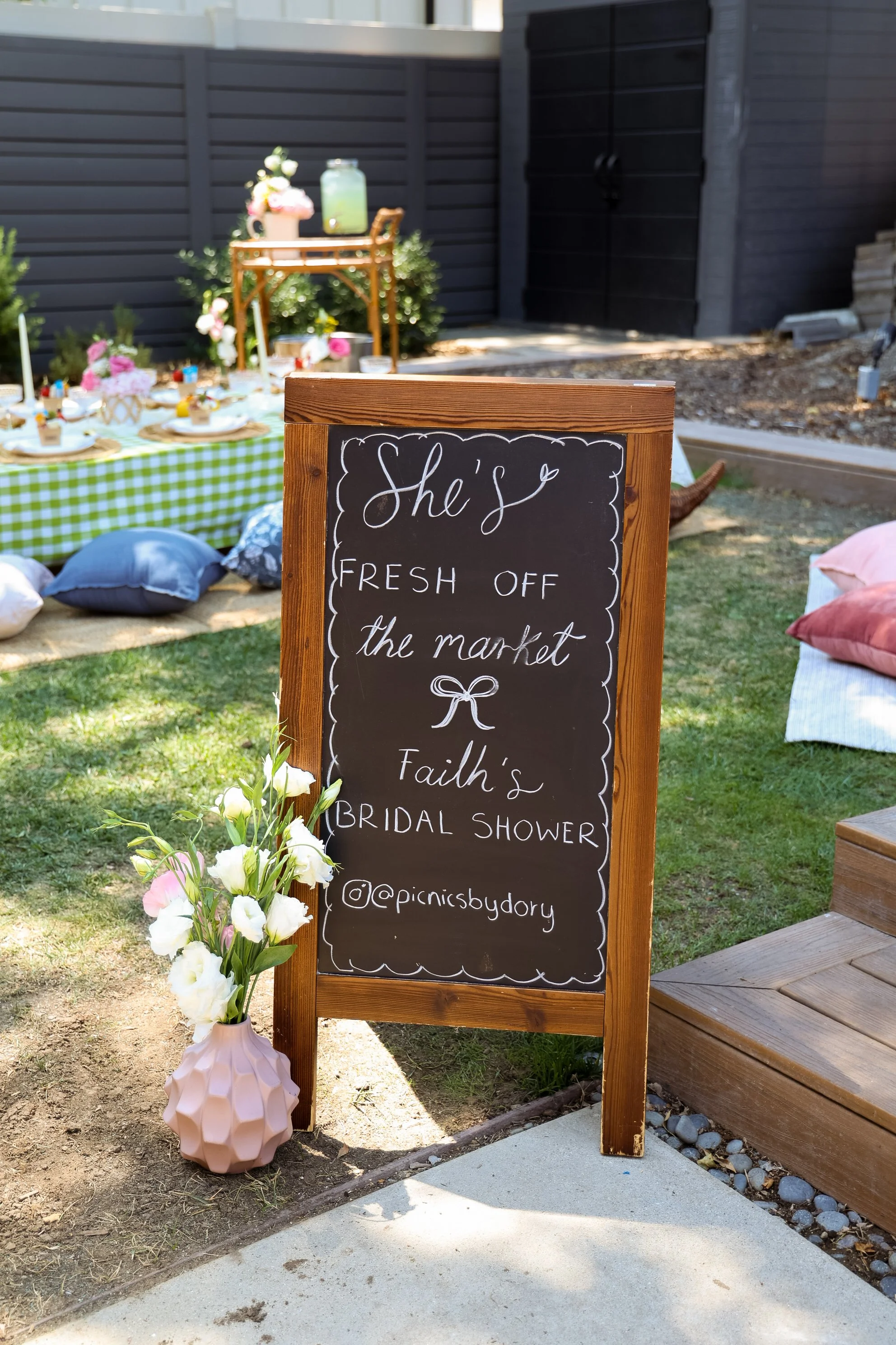 A chalkboard sign with decorative border and handwritten message announcing a bridal shower for Faith, located outdoors near a table with pillows and picnic setting, and decorated with flowers in a pink vase.