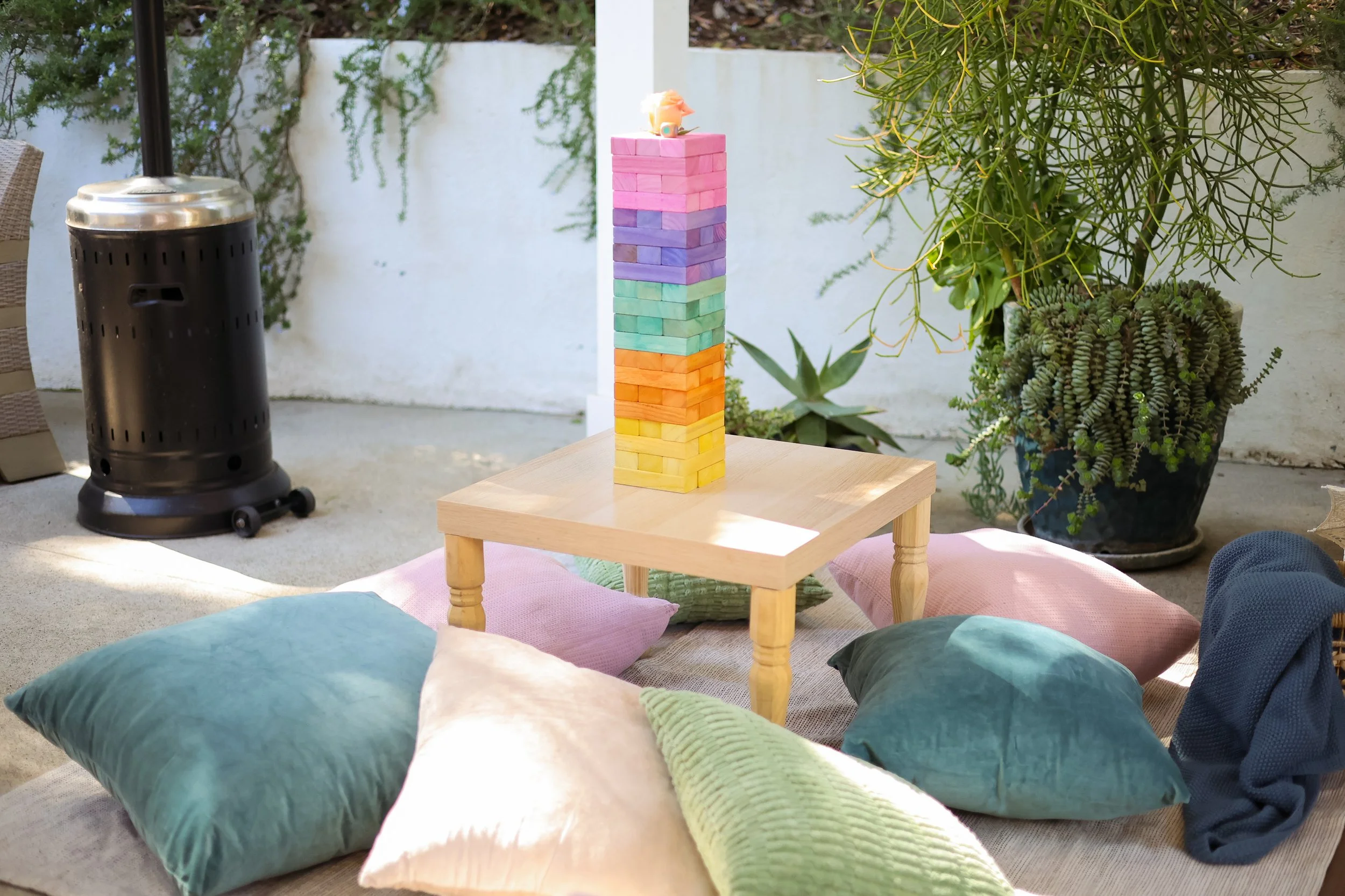 Outdoor patio scene with a small wooden table surrounded by colorful cushions on the ground. On the table is a tower of pastel-colored Jenga blocks. To the left is a black patio heater, and to the right are potted green plants, including succulents, against a white wall.