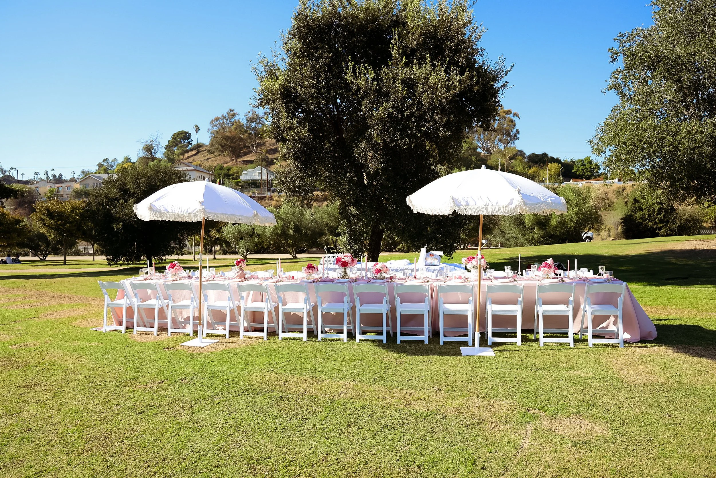 Outdoor wedding or event table setup on a grassy field with pink flowers, white chairs, and large white umbrellas, surrounded by trees and a hilly background.