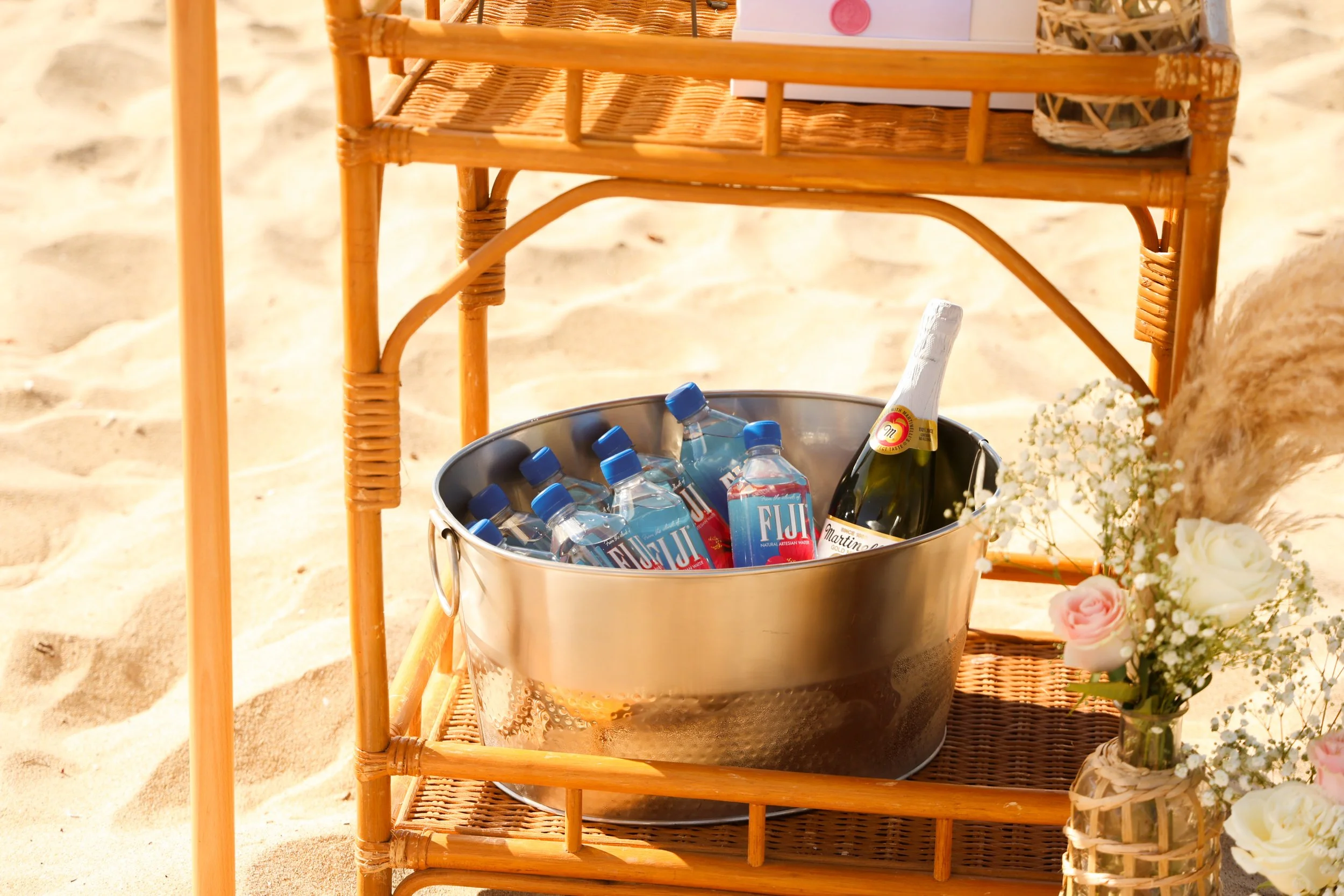 A metal bucket containing bottled water and a bottle of sparkling wine on a wicker side table on a sandy beach, with a small vase of flowers nearby.