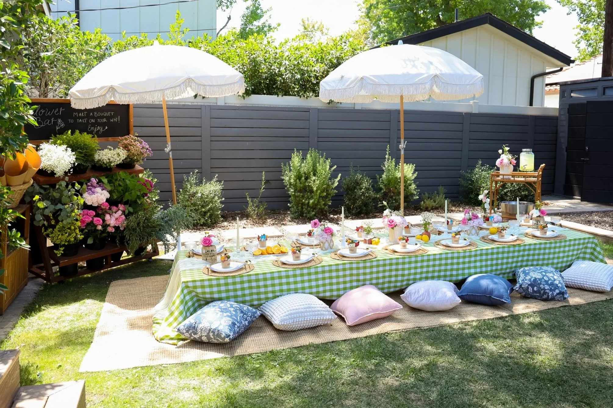Outdoor setting with a long picnic table covered with a green and white checkered tablecloth, decorated with small flower arrangements, lit candles, and pastel-colored desserts. Surrounding the table are cushions on a woven mat. Two white umbrellas provide shade. To the left, there is a display of potted flowers and a chalkboard sign that reads "Make a bouquet to take on your way! Enjoy." A small wooden cart with more floral arrangements is on the right.