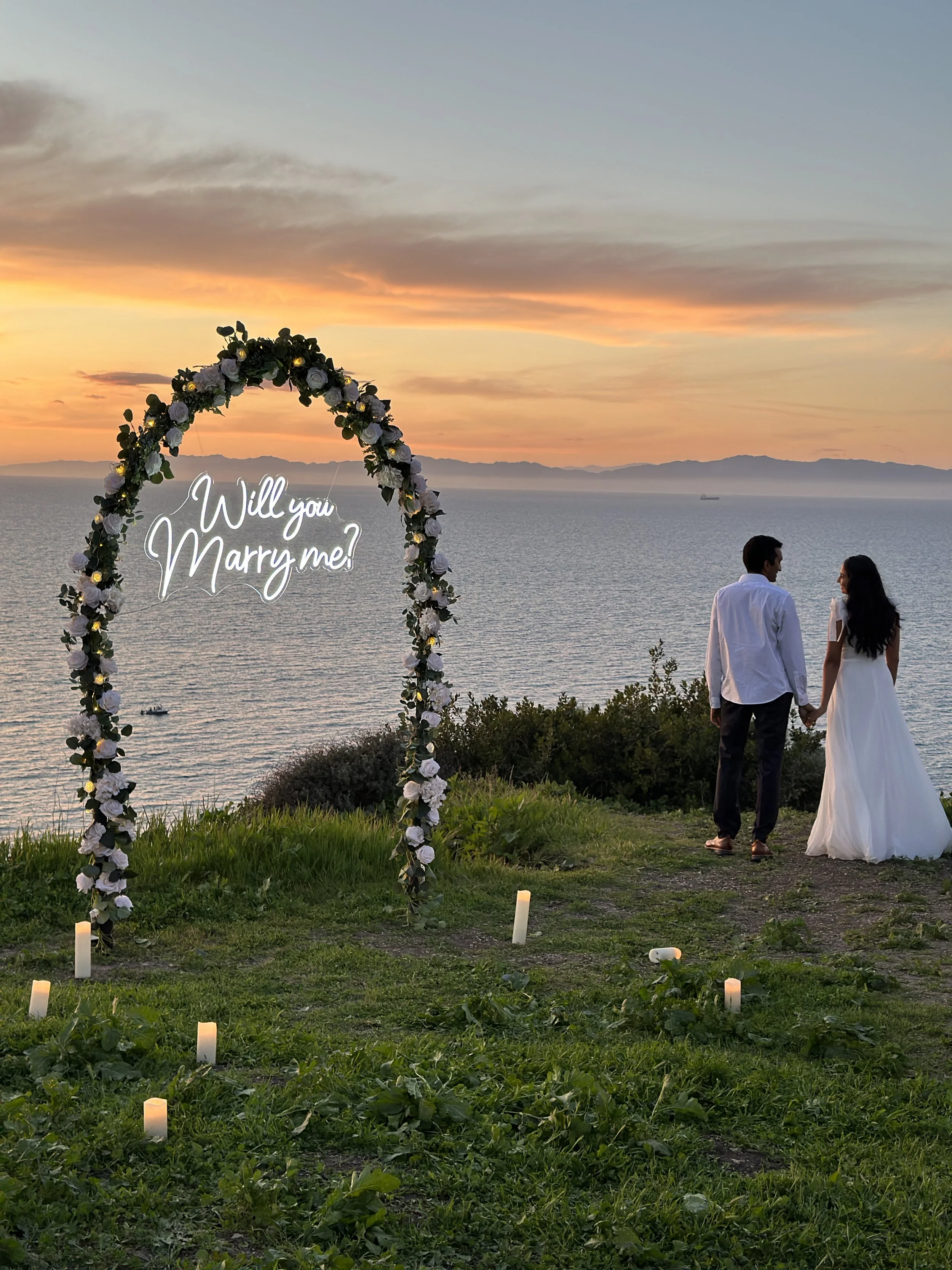 A couple holding hands at sunset near a lake, with a floral arch that says "Will you Marry me?" and candles on the grass.