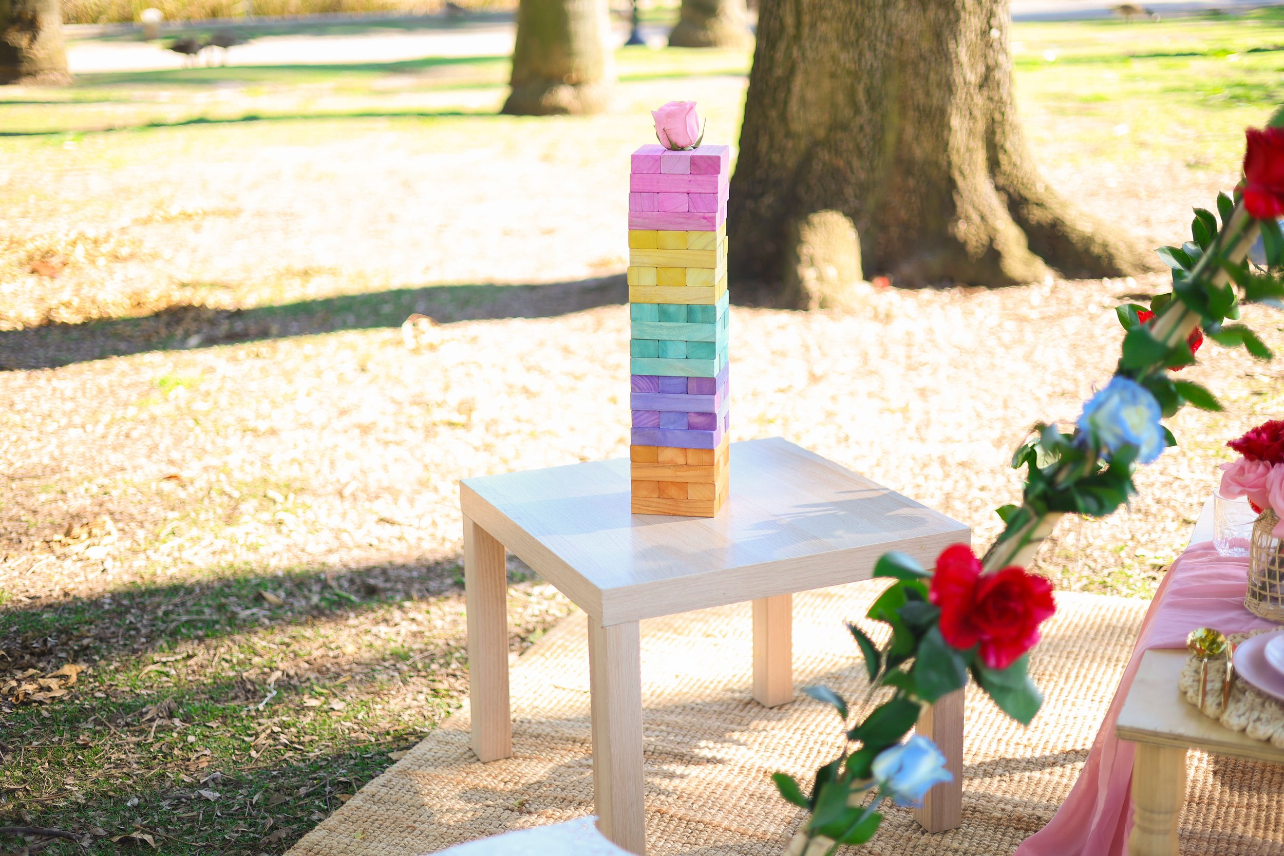 Colorful wooden blocks stacked into a tower on a small white table outdoors, with garden and trees in the background.