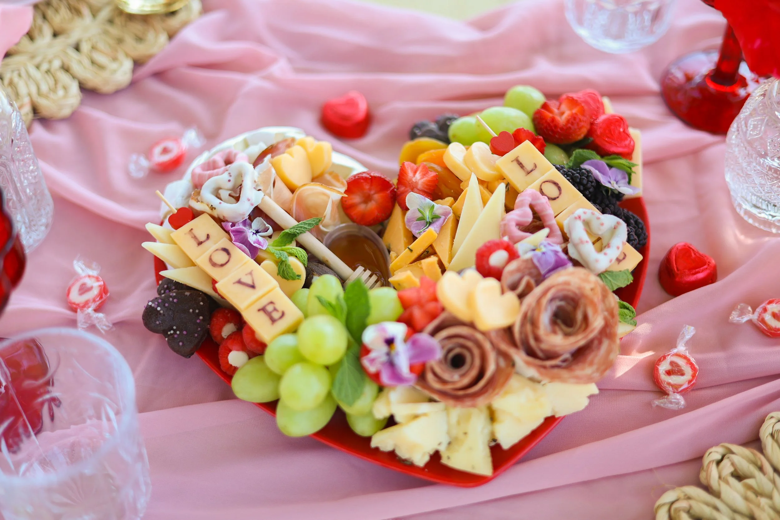 Heart-shaped platter of assorted cheeses, grapes, strawberries, blackberries, and edible flowers on a pink tablecloth.