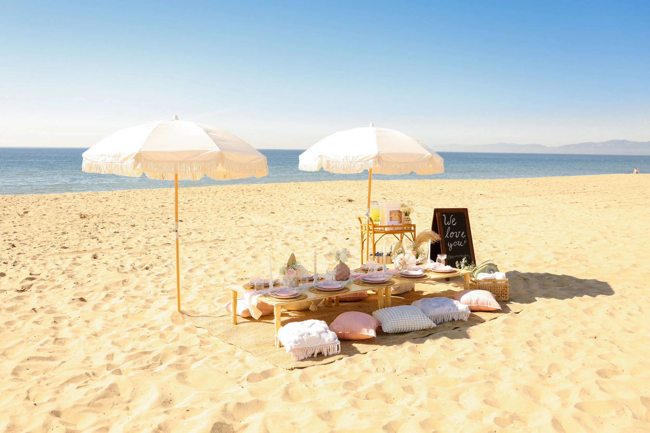 Beach picnic setup with two white umbrellas, a low table with pink plates, flowers, and candles, surrounded by cushions on the sand, near the ocean.