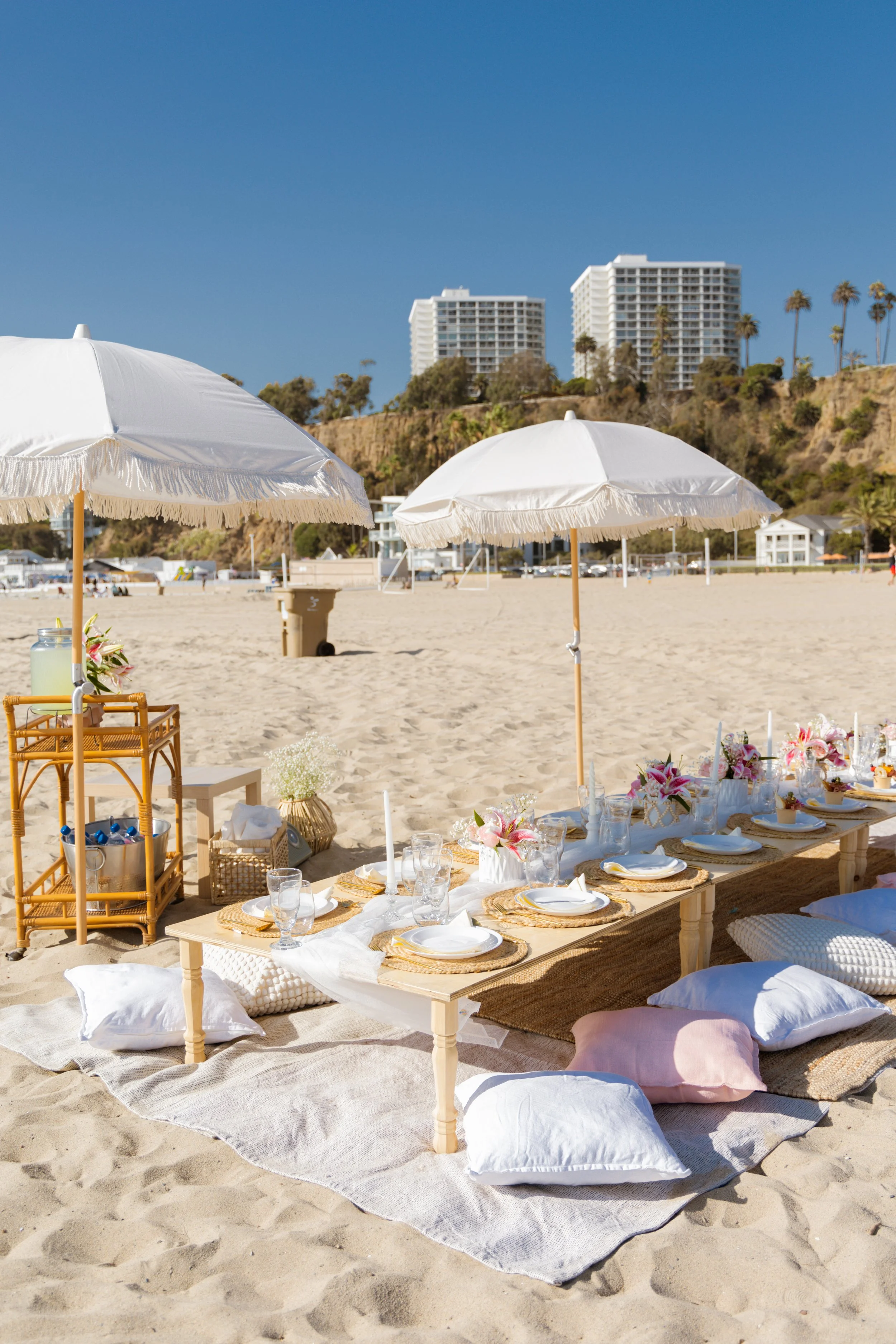 Beachside dining setup with a low wooden table, cushions, white plates, glasses, and pink flowers under umbrellas, with high-rise buildings, palm trees, and a cliff in the background.