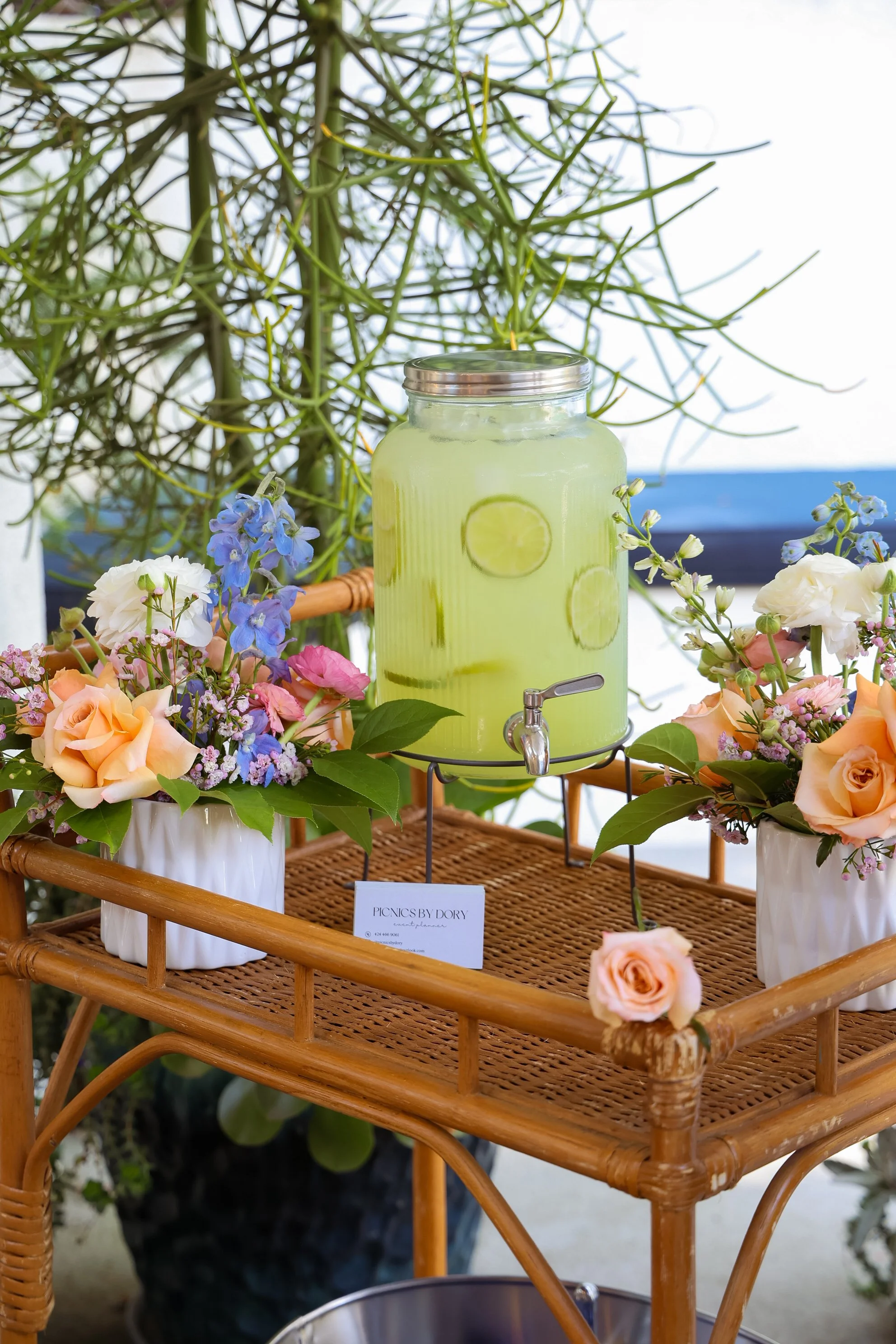 A large glass beverage dispenser filled with lemonade containing lemon slices, placed on a wicker cart decorated with peach and white roses and blue flowers.