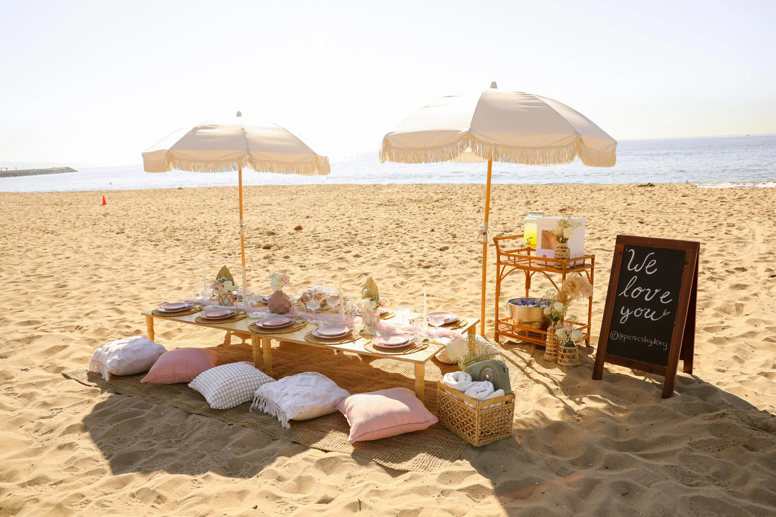 Beach picnic setup with a low wooden table, pink and white pillows, two white umbrellas, and a chalkboard sign that reads "We love you". The table is decorated with plates, candles, and small floral arrangements, set on sand near the ocean.