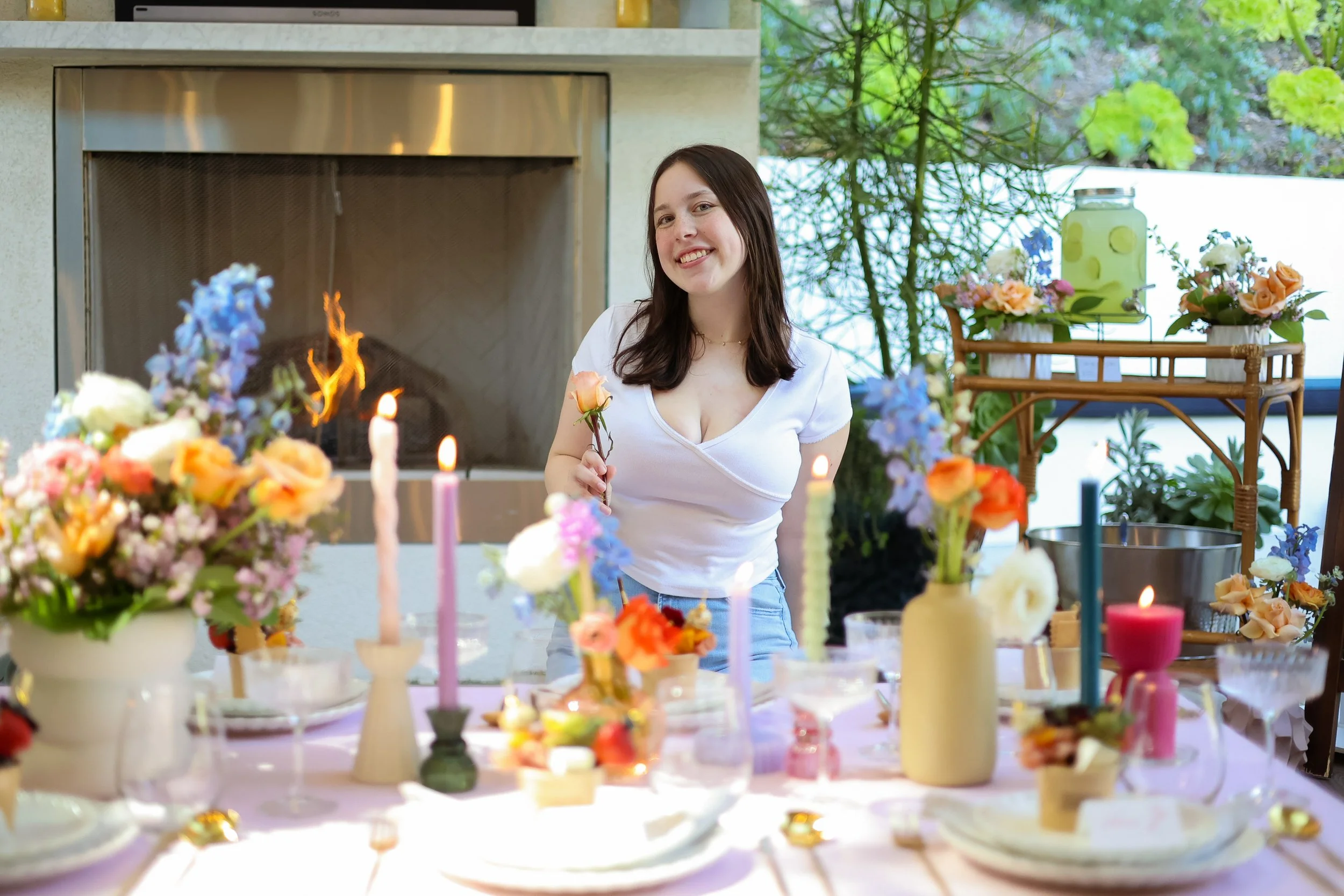 A woman with dark brown hair smiling at a table decorated with colorful flowers, candles, and tableware, standing near a fireplace with a green garden visible through the window.