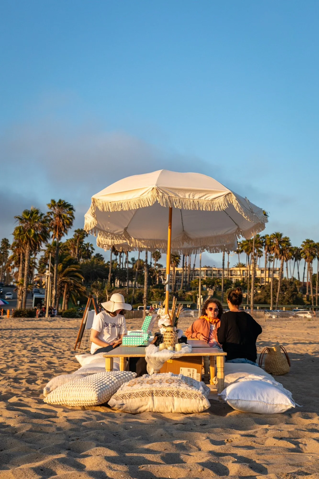 Three women sitting on the beach under a large white beach umbrella, surrounded by blankets and pillows, with palm trees and buildings in the background during sunset.