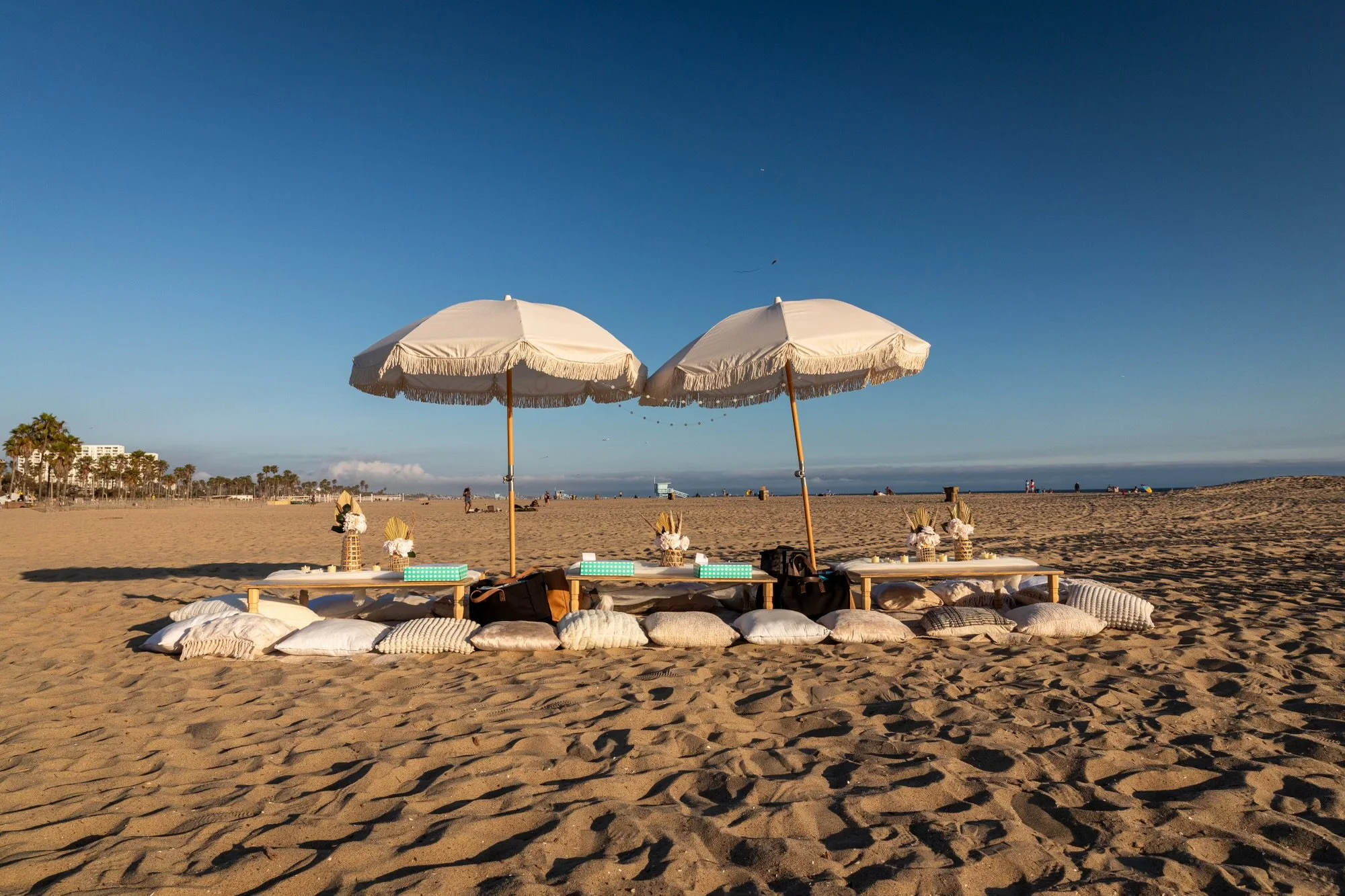 Beach with two white umbrellas, low tables, and pillows arranged on sand, with calm ocean and distant palm trees and buildings in the background.