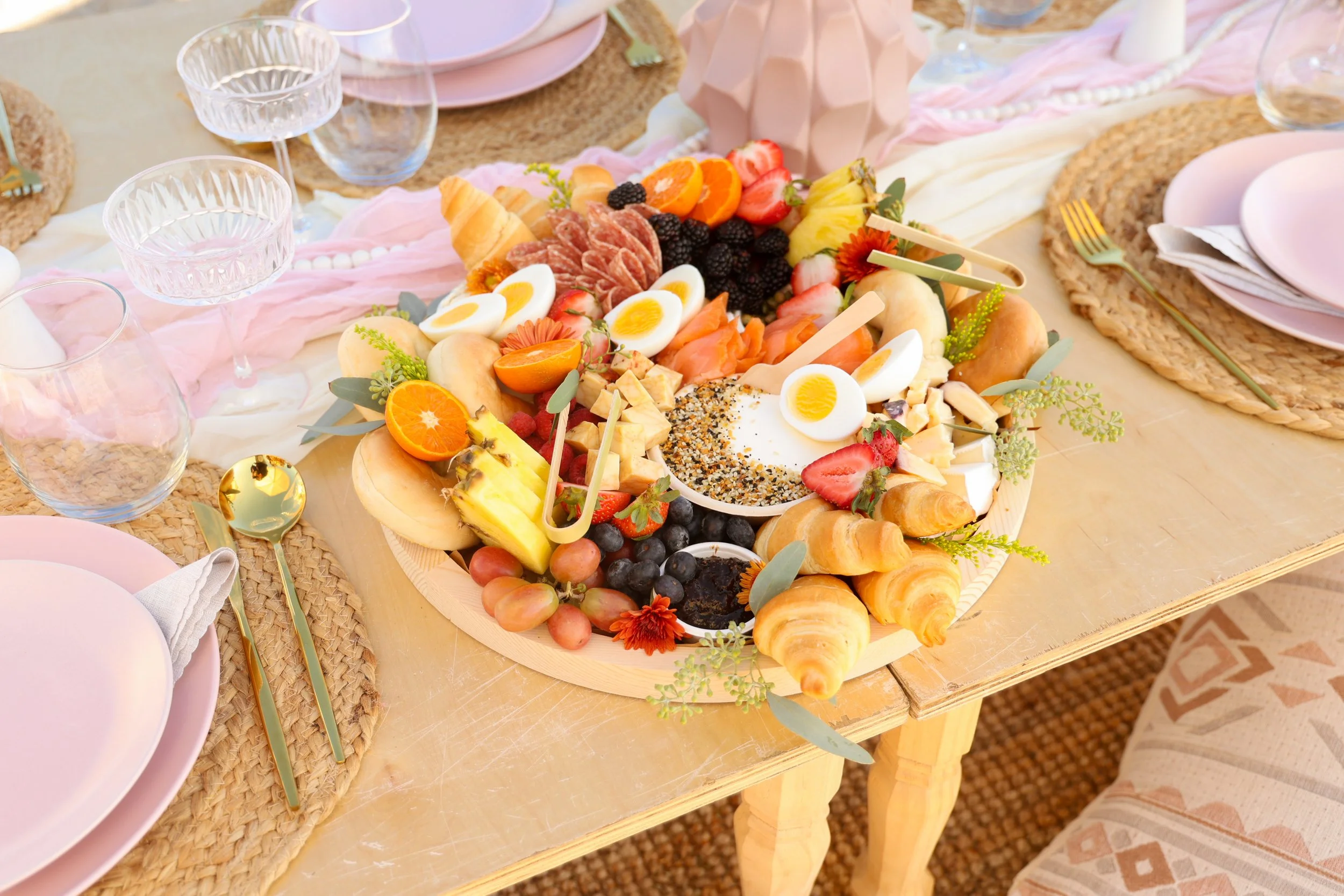 A wooden table decorated with a pink cloth and a large round wooden serving tray filled with an assortment of fruits, cheeses, and pastries, surrounded by pink plates, gold utensils, and empty glasses for a festive gathering.