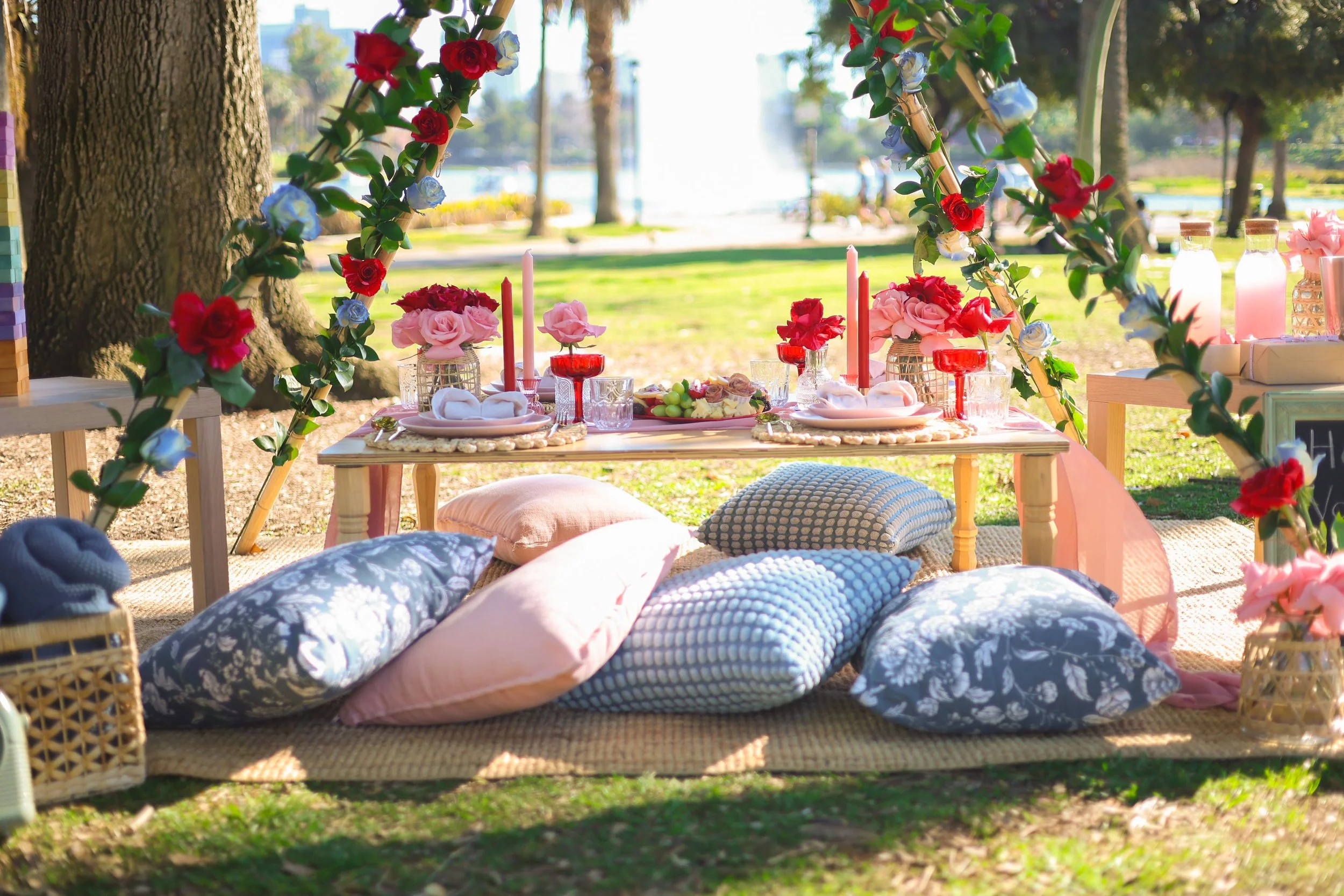 Decorative outdoor picnic setup with a low table, floral arrangements, colorful cushions, and a floral arch, set in a park near water.