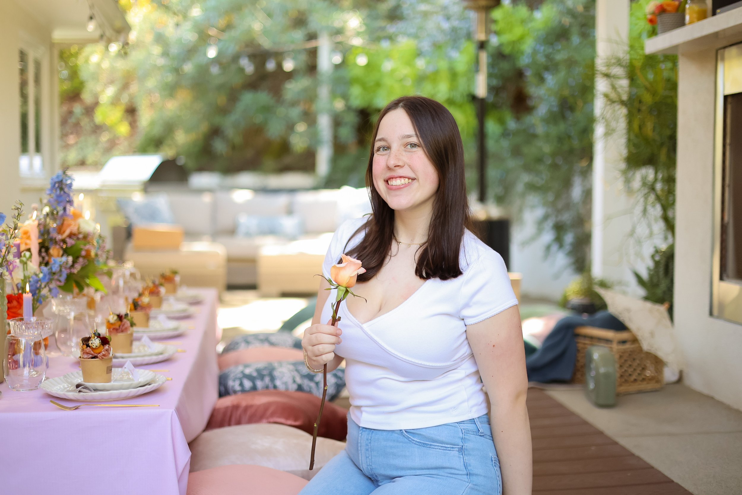 A young woman with dark brown hair smiling and holding a pink rose in her hand, standing outdoors at a decorated table with food and flowers, with a garden and sofa in the background.