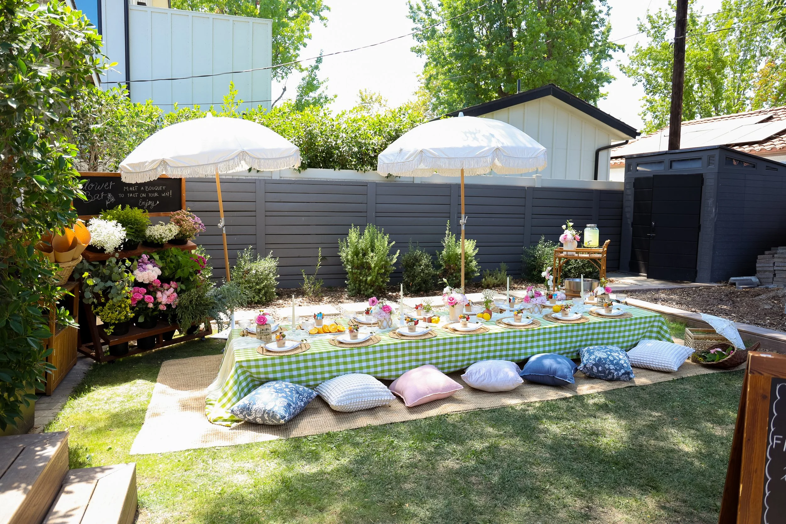 An outdoor garden setting for a gathering, featuring a long table with a green and white checkered tablecloth, surrounded by cushions on the grass. Two white umbrellas provide shade, and the table is decorated with flowers, plates, and candles. There