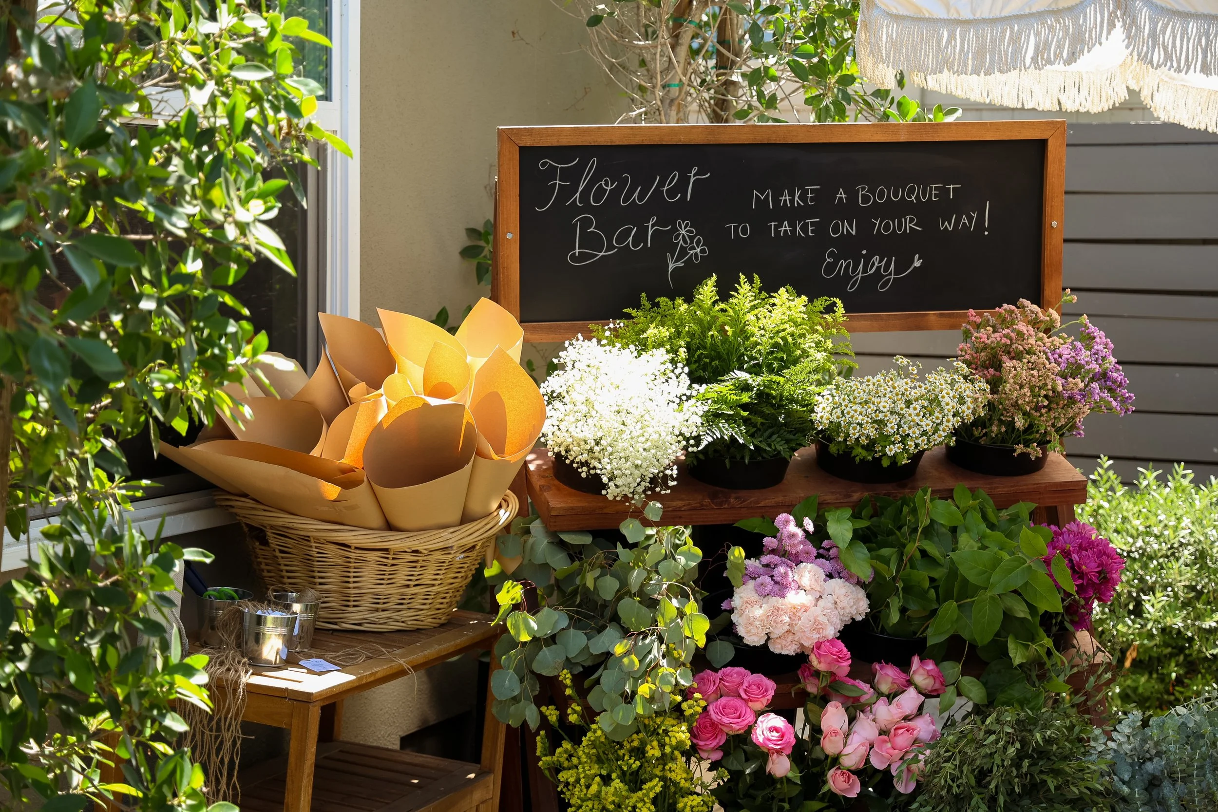 A flower stand outside with potted flowers and a blackboard sign that reads 'Flower Bar, Make a bouquet to take on your way! Enjoy.' A basket with paper cone sleeves for wrapping flowers is nearby.