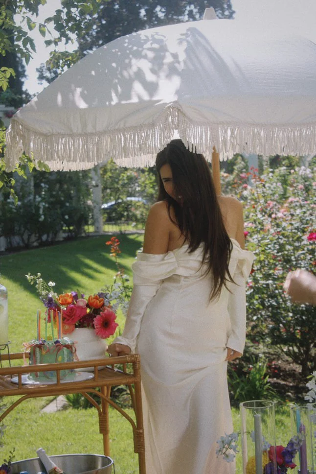 A woman in a white off-shoulder dress standing outdoors under a white parasol with fringes, next to a table with a birthday cake, flowers, and colorful candles in a garden setting.