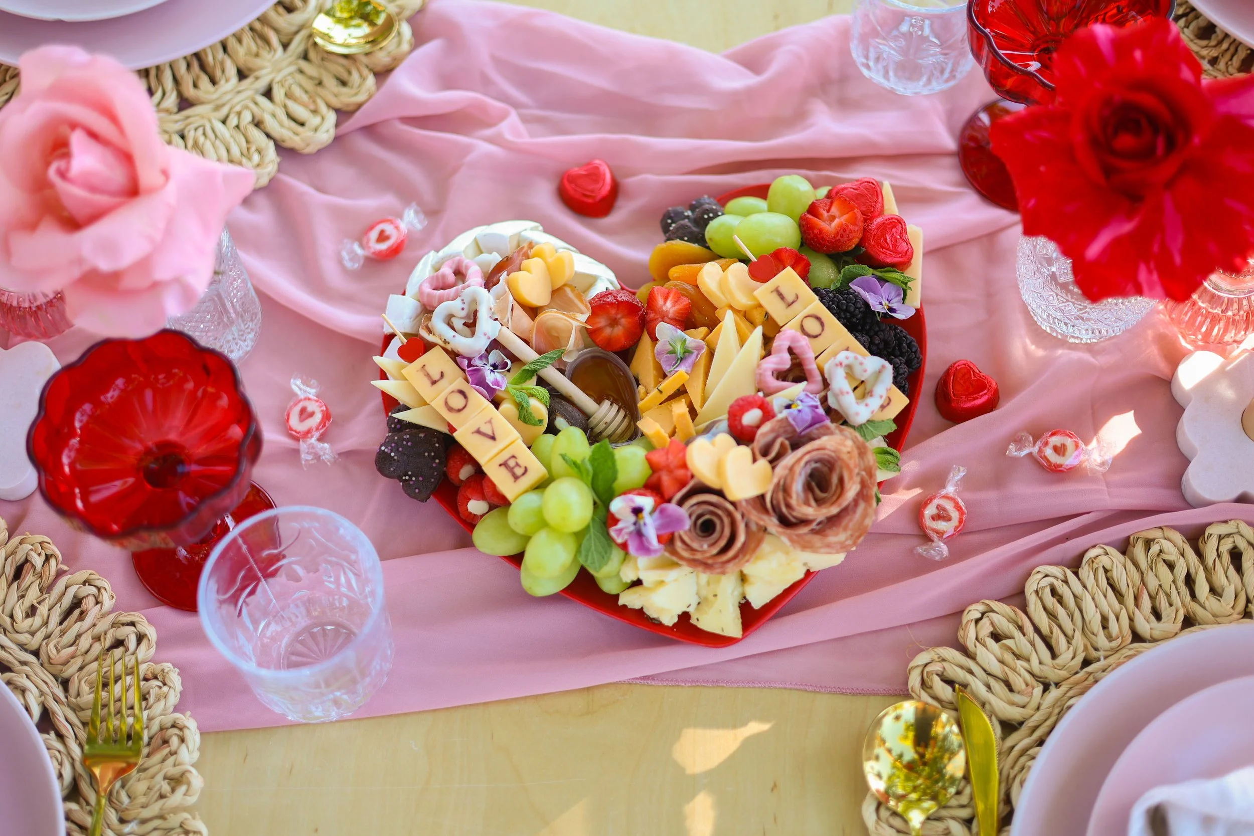 Heart-shaped cheese and fruit platter with grapes, strawberries, blackberries, cheese, chocolates decorated with "LOVE" blocks, surrounded by flowers, candy, glasses, and tableware on a pink cloth.