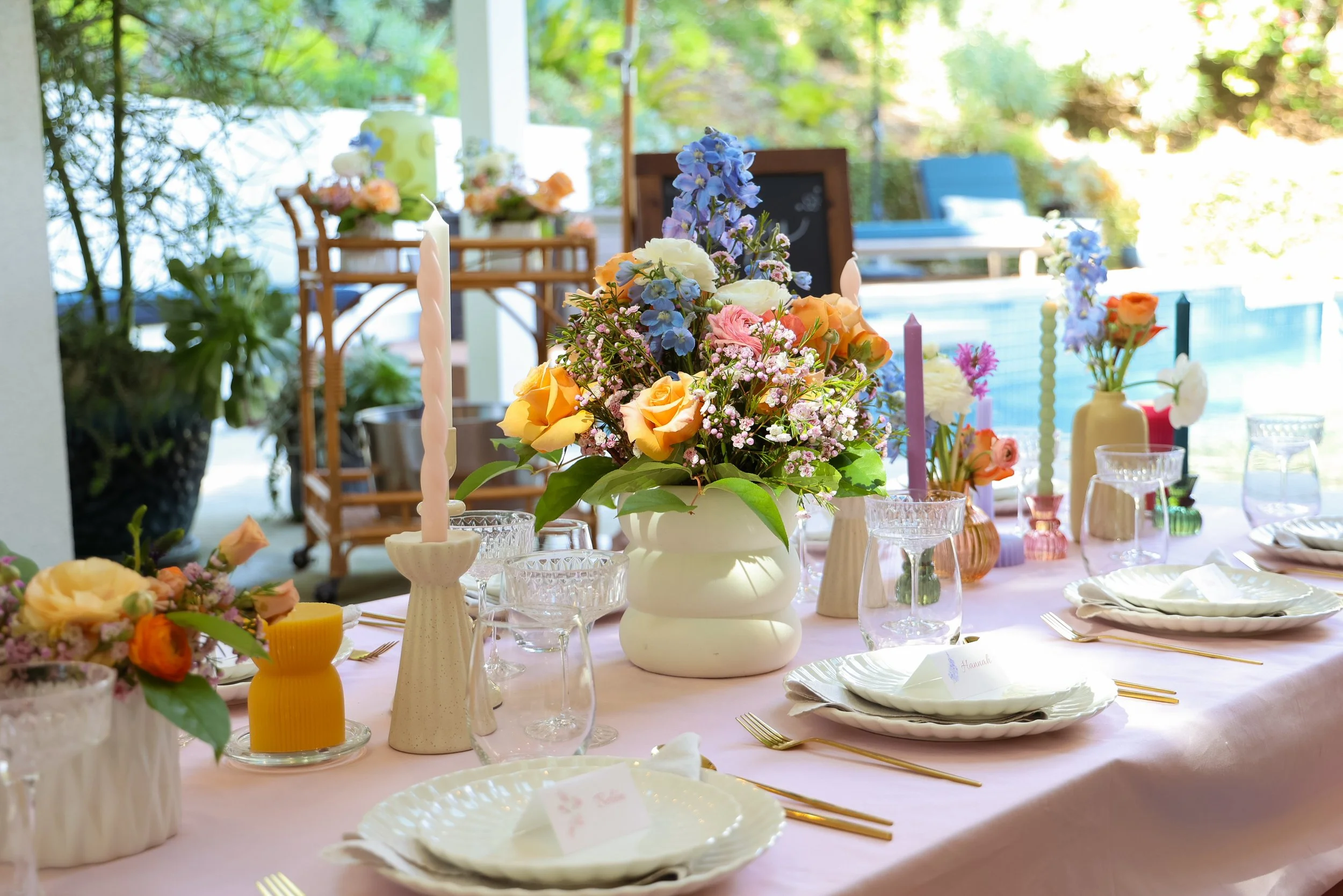 A table set for a celebration with floral centerpieces in vases, candles, glassware, plates, and gold-toned cutlery, outdoors near a pool.