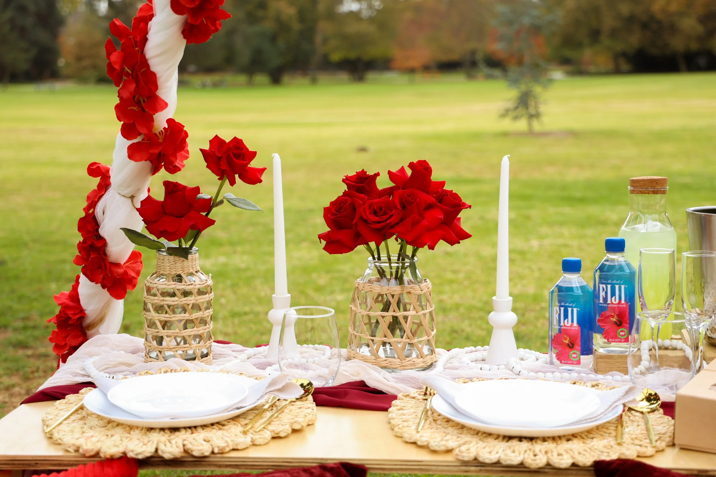 Table set outdoors with white plates, gold utensils, wine glasses, and floral arrangements with red roses in wicker vases, surrounded by white candles and Fiji water bottles, with a grassy field and trees in the background.