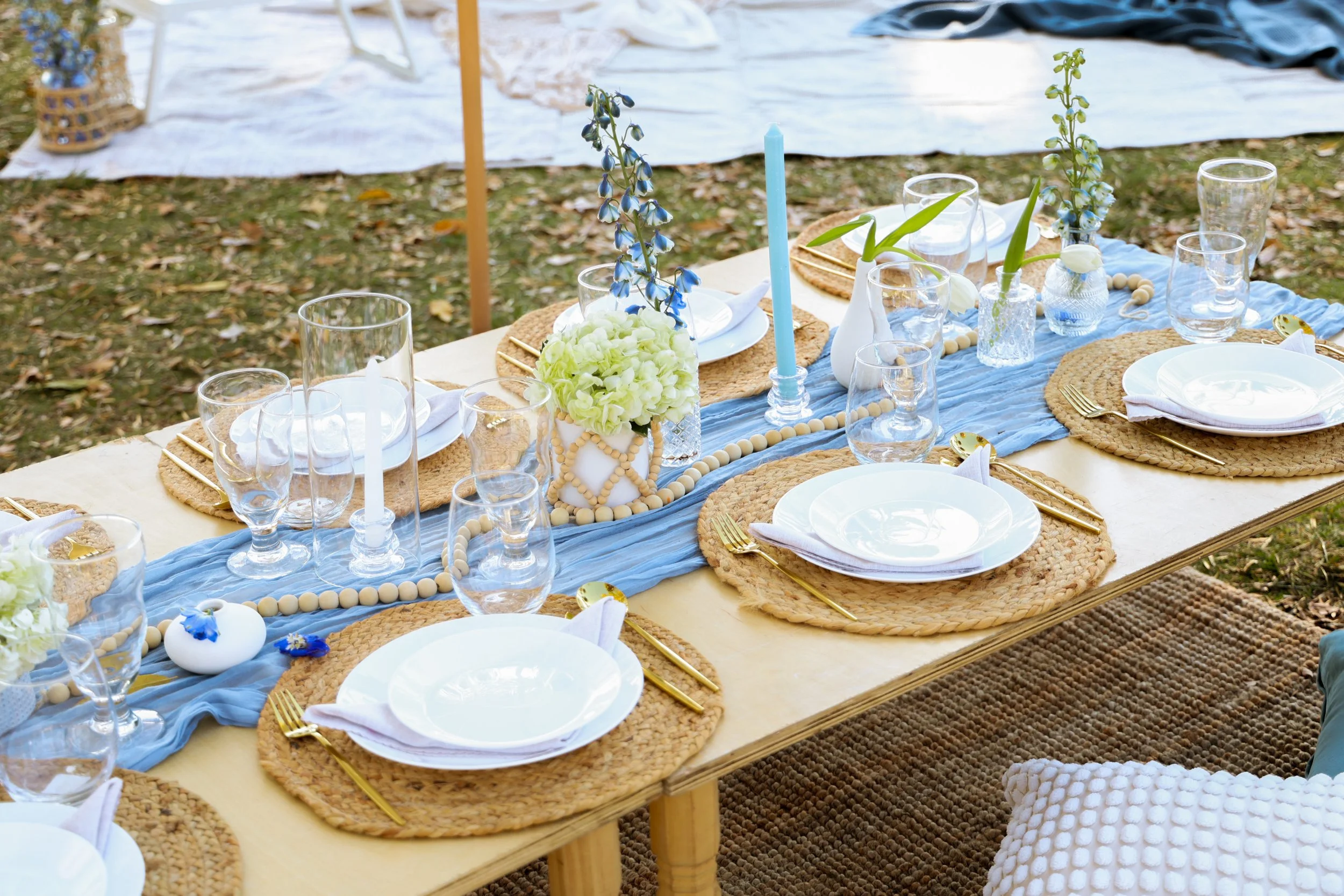 Outdoor dining table decorated with white and blue flowers, candles, and tableware, set for a meal.