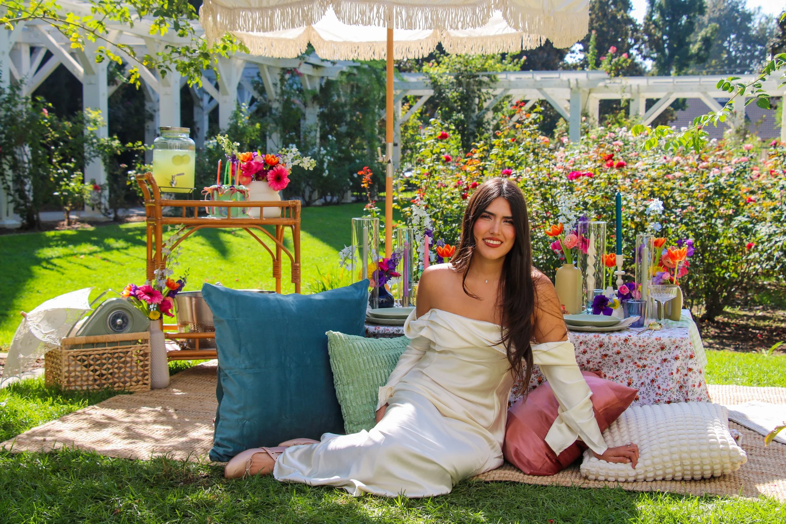 A young woman with long dark hair and a white off-the-shoulder dress sitting on a picnic blanket at a garden party, surrounded by colorful pillows, flowers, and table settings under a large cream-colored umbrella.