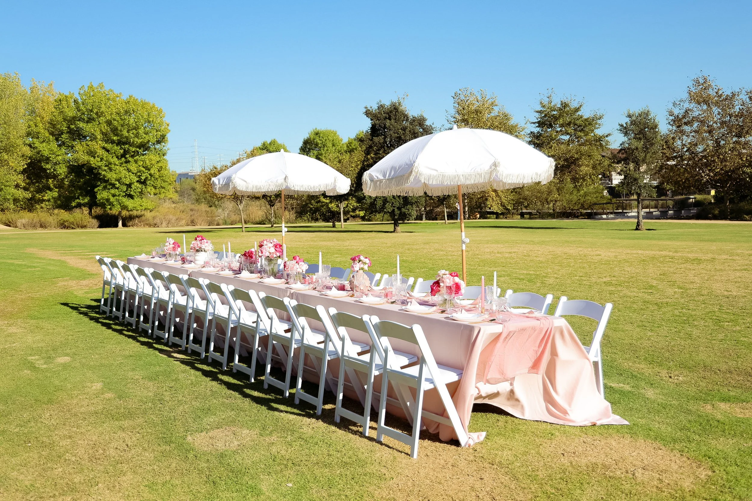 Outdoor banquet table set with pink flowers, candle holders, and tableware, shaded by two large white umbrellas in a grassy park with trees in the background.