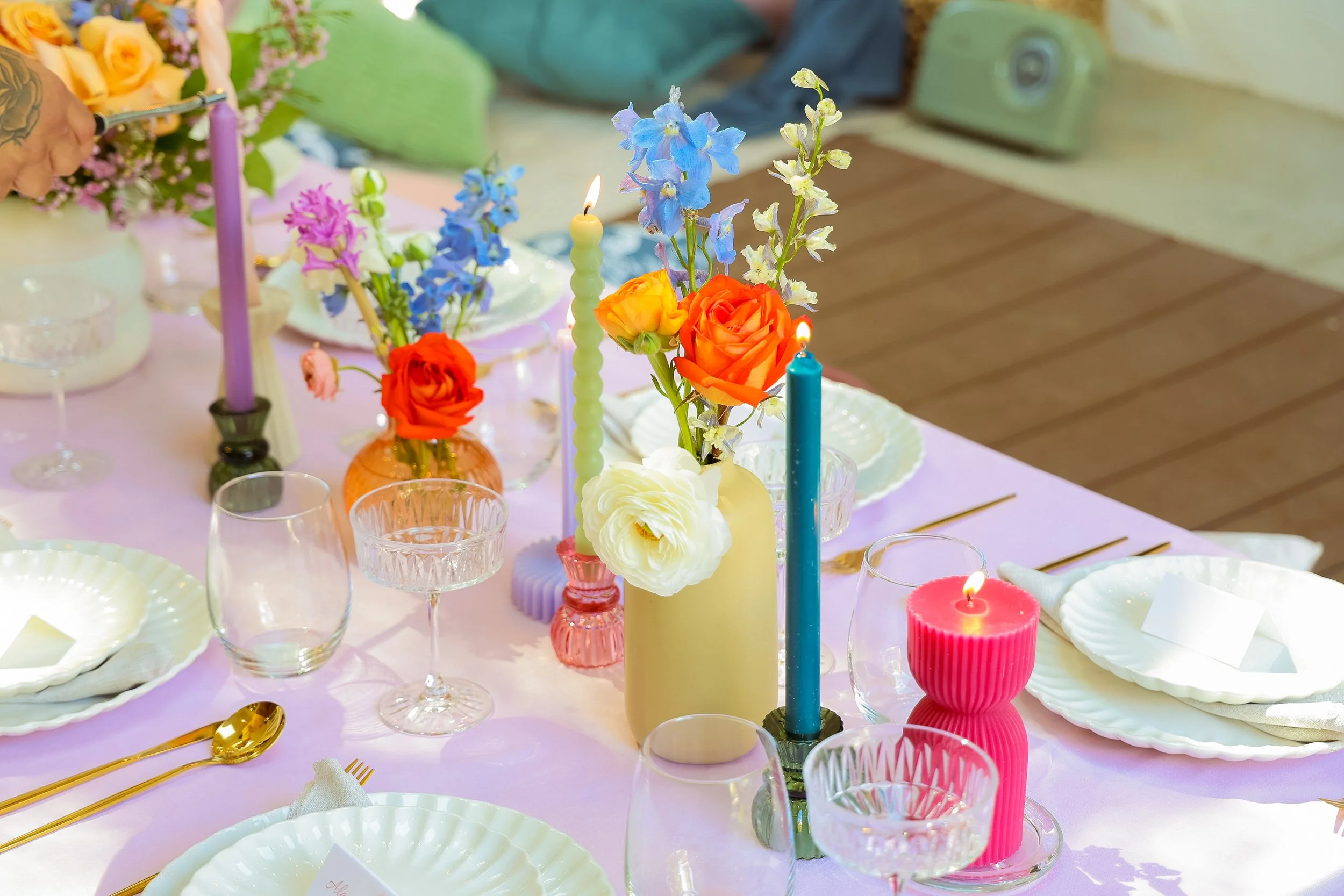 A pastel-colored table setting with floral centerpieces, striped candles, plates, glasses, and gold utensils, decorated for a celebration.