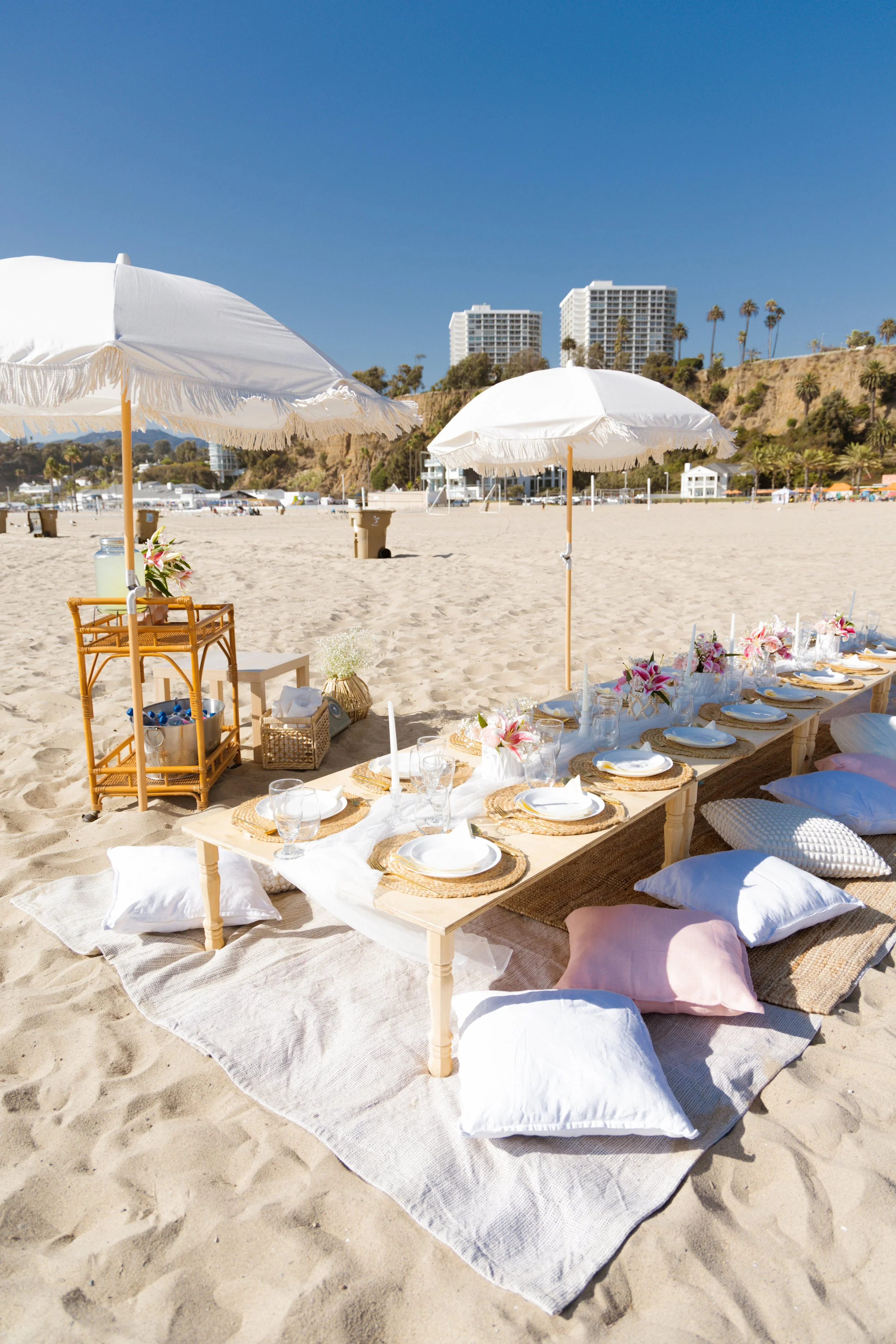 Beachside outdoor dining setup with a low table, cushions, white umbrellas, and floral decorations on sandy beach with buildings and palm trees in the background.