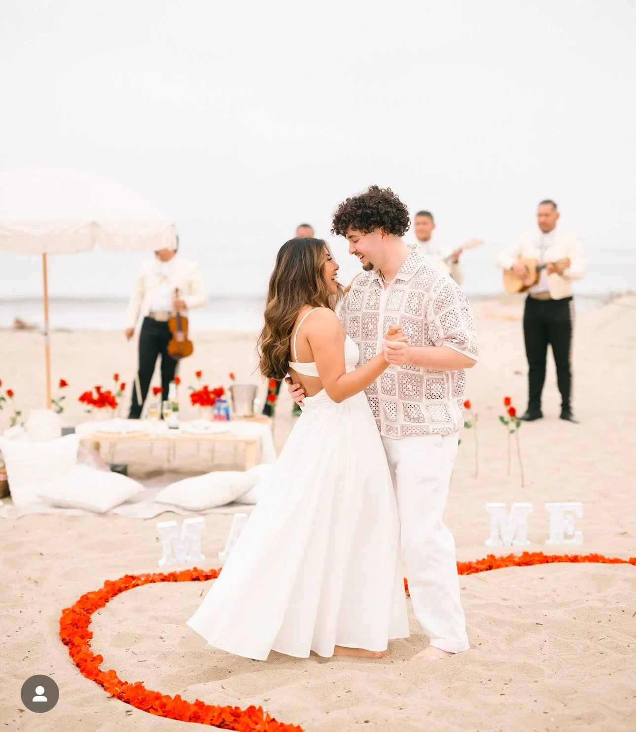 A couple dancing on a beach during their wedding, with a heart-shaped flower arrangement around them and a band playing in the background.
