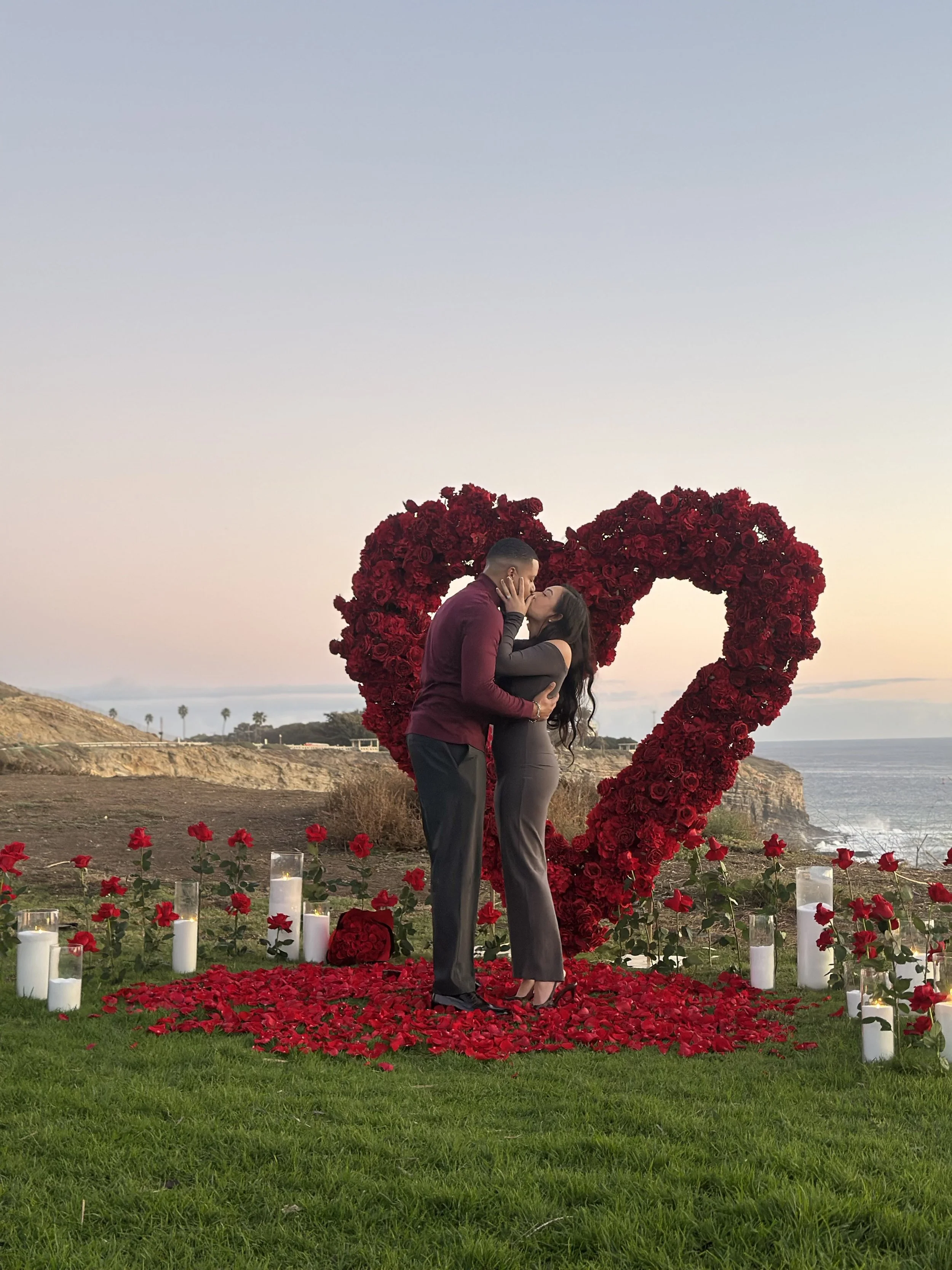 A couple sharing a kiss in front of a heart-shaped floral display made of red roses, with candles and rose petals on the ground by the ocean at sunset.