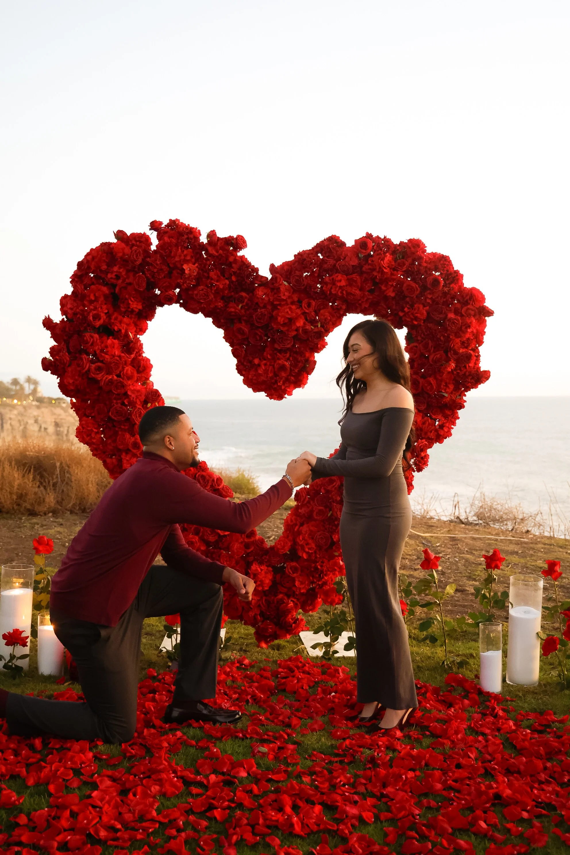 A man proposing marriage to a woman outdoors near the ocean, with a large heart-shaped flower arrangement in the background, surrounded by red roses and candles.