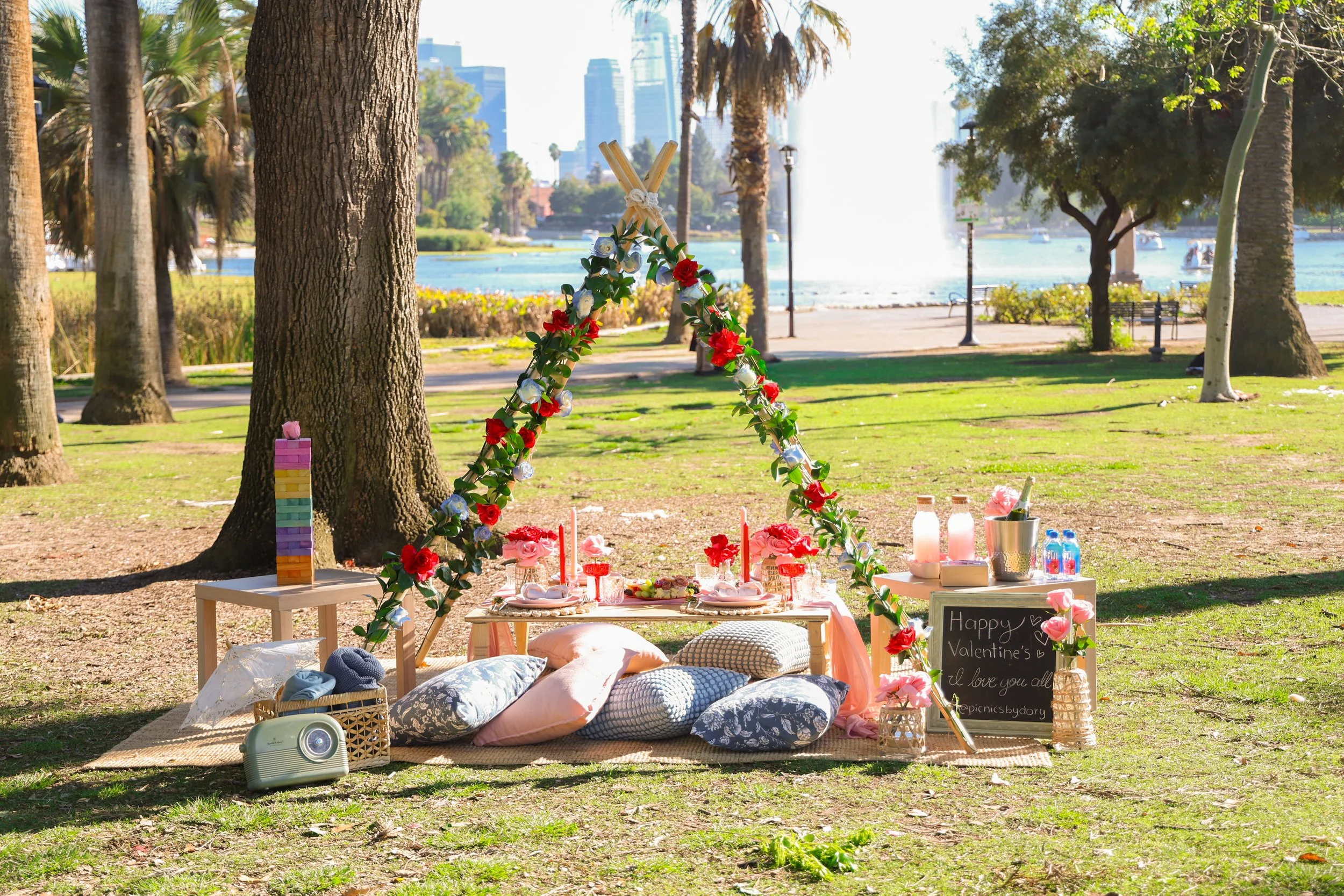 A decorated outdoor picnic setup near a lake with trees, a fountain, and city skyline in the background. The setup includes pillows, a small table with pink flowers, candles, drinks, and a chalkboard sign wishing a happy Valentine's Day.