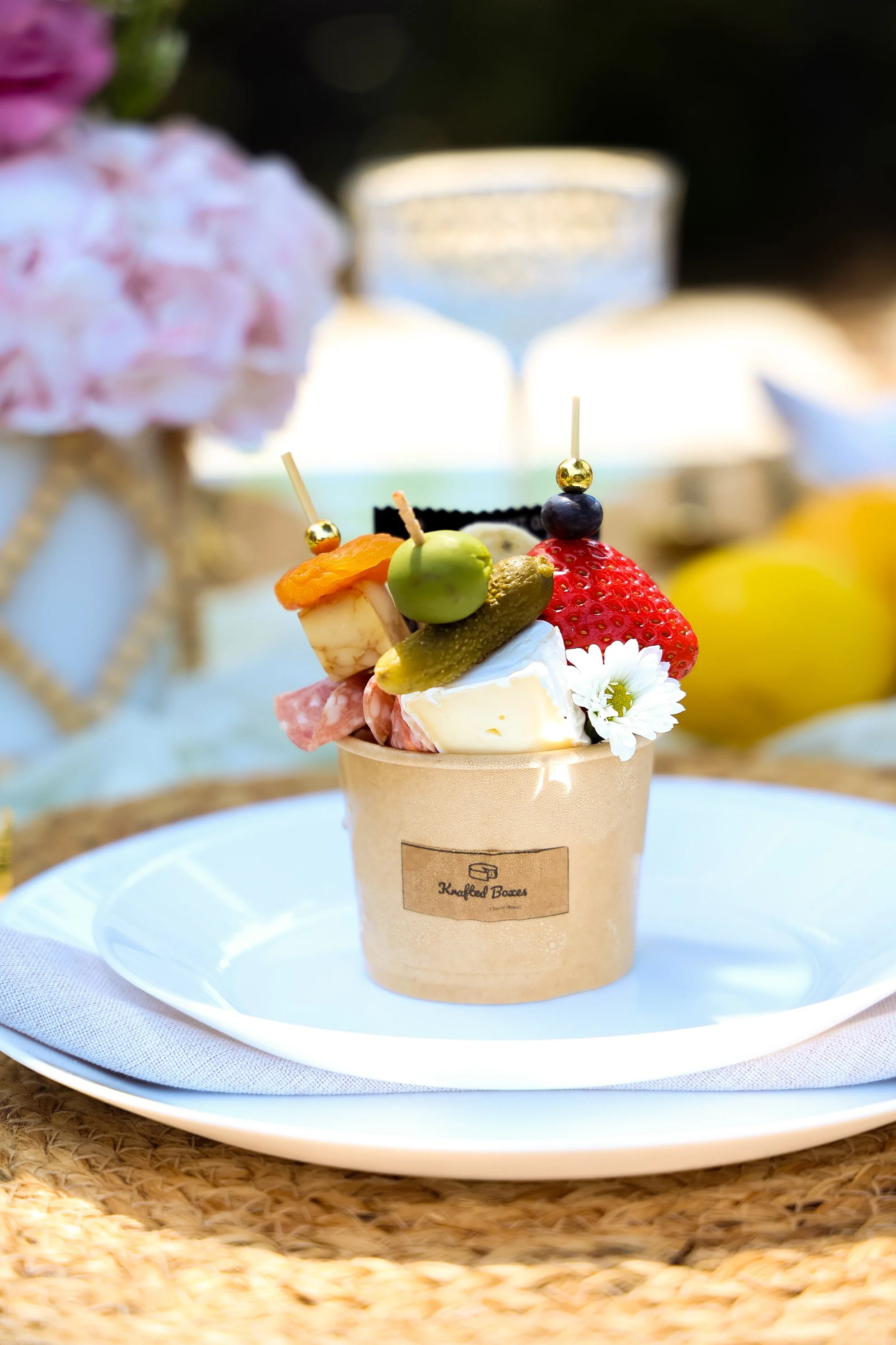 A cup filled with various fresh fruits including strawberries, grapes, an apple, a pickle, and a cheese cube, garnished with a small white flower, placed on a white plate with a light blue napkin, set on a woven placemat.