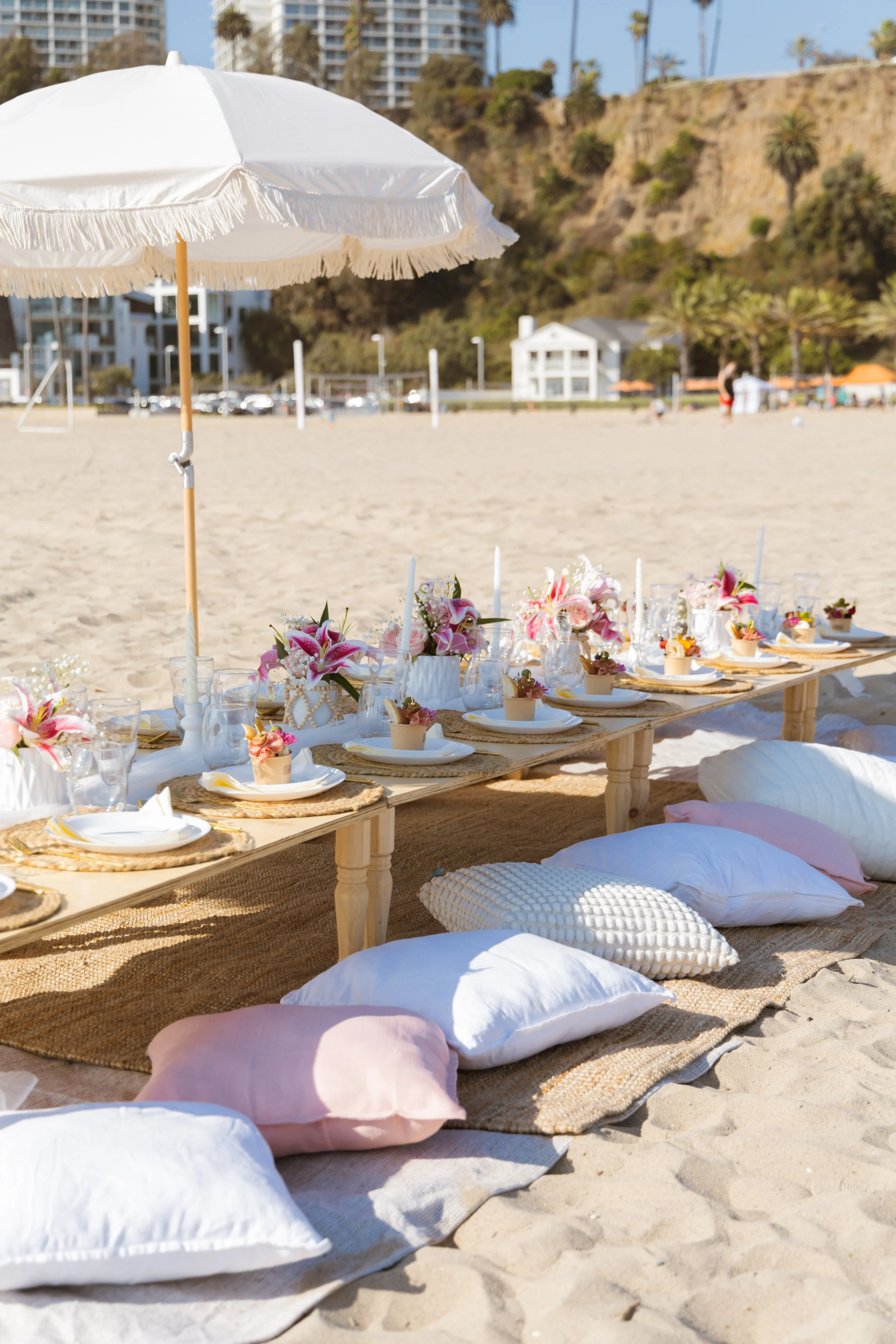 A beach setting with a low wooden table decorated with pink and white flowers, surrounded by cushions and pillows on the sand. There is a large white umbrella providing shade, with a background of palm trees, buildings, and a hillside.