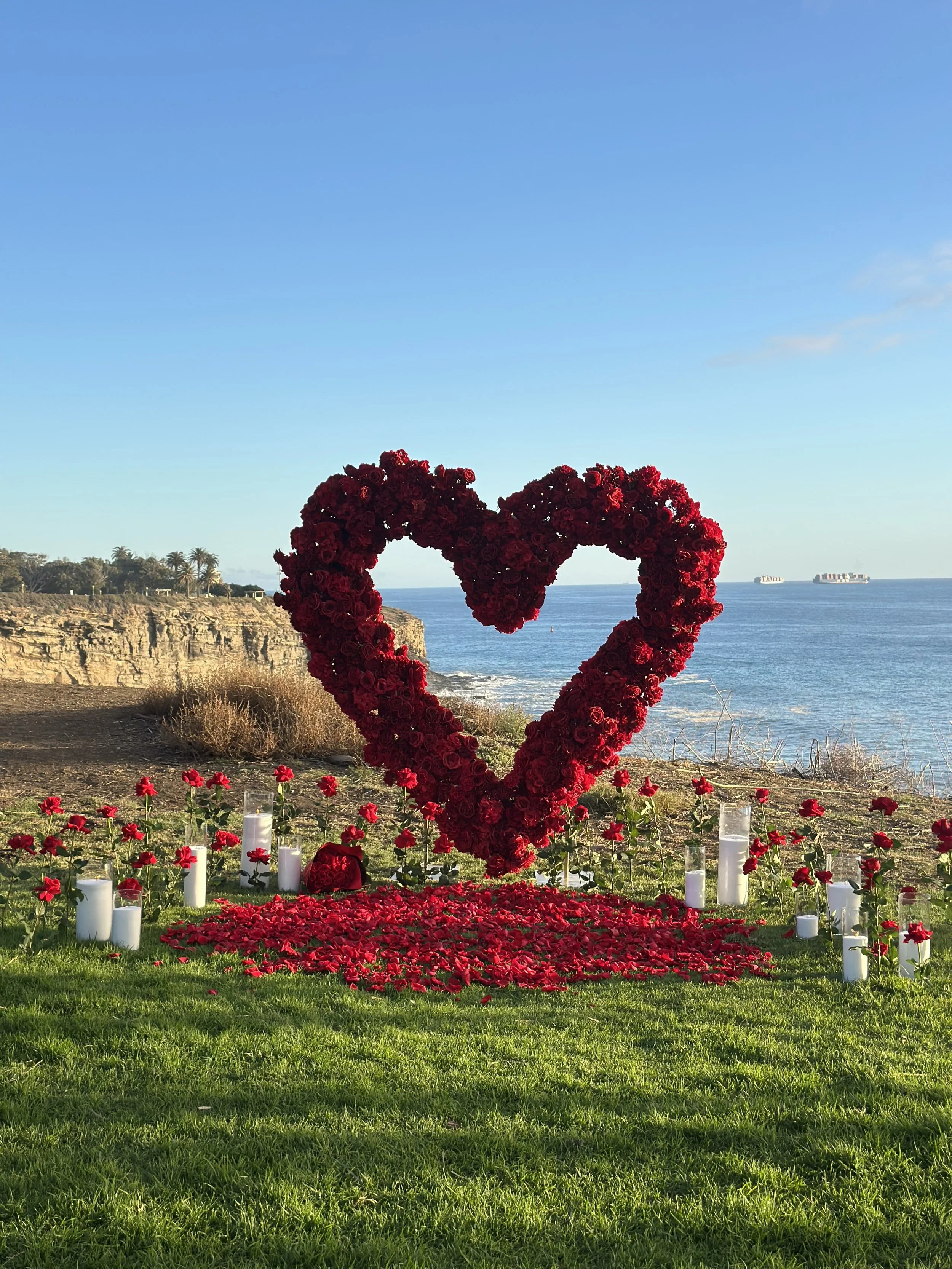 A large heart-shaped structure made of red roses by the ocean, surrounded by candles and rose petals on the grass.