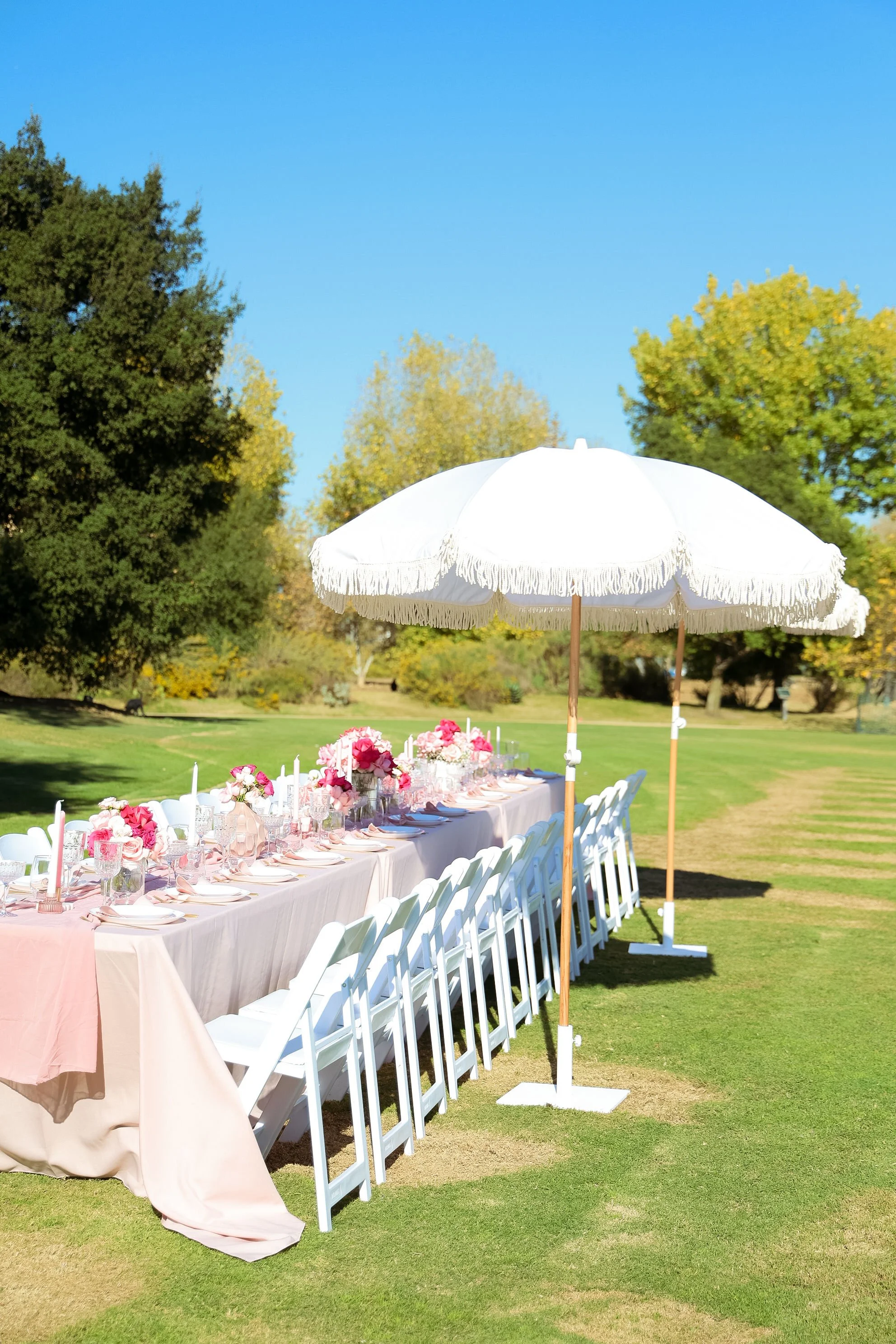 Outdoor event table setup on a grassy lawn with pink and white floral centerpieces, candles, and white chairs, shaded by large white umbrellas under a clear blue sky.