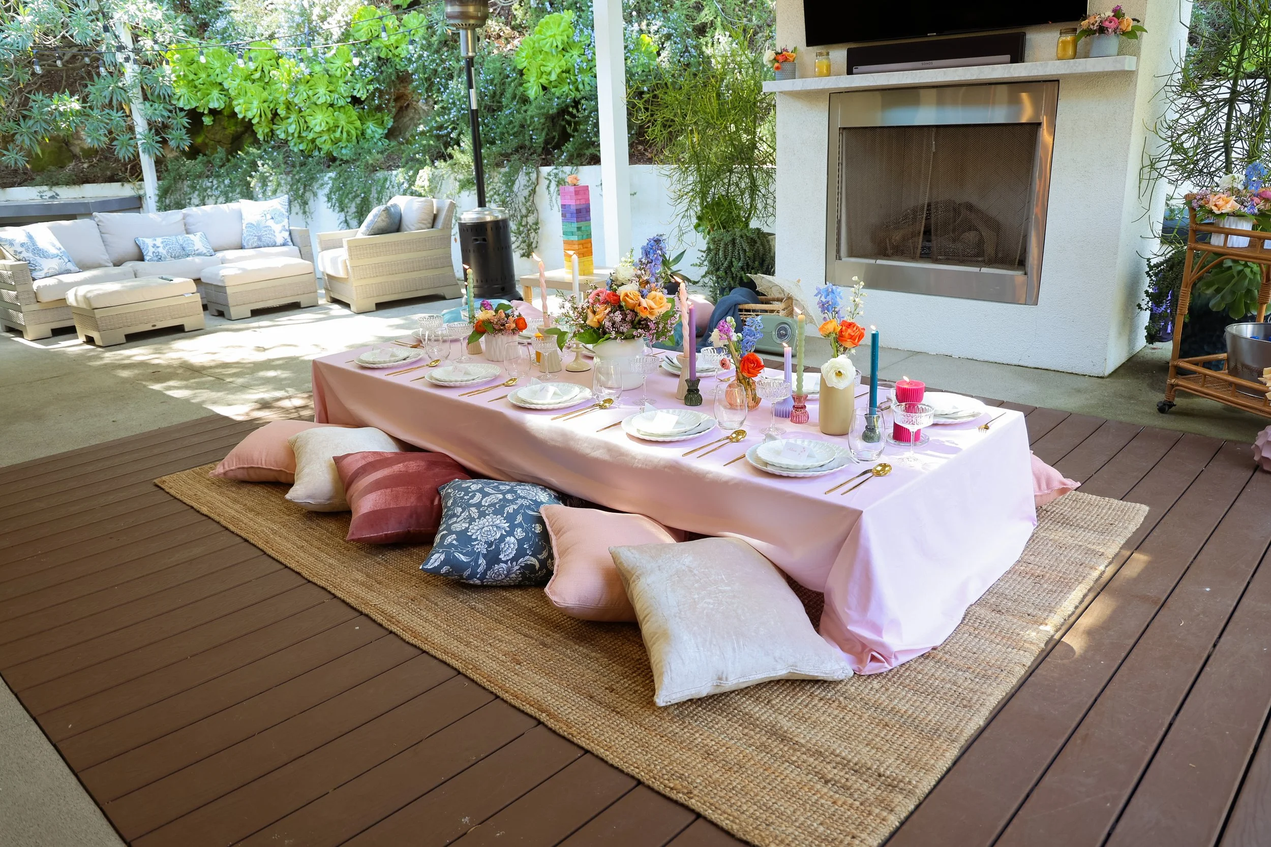 Outdoor patio decorated for a gathering with a long table draped in pink cloth, surrounded by colorful pillows on a woven rug, with floral centerpieces and candles, in front of a white fireplace and greenery.
