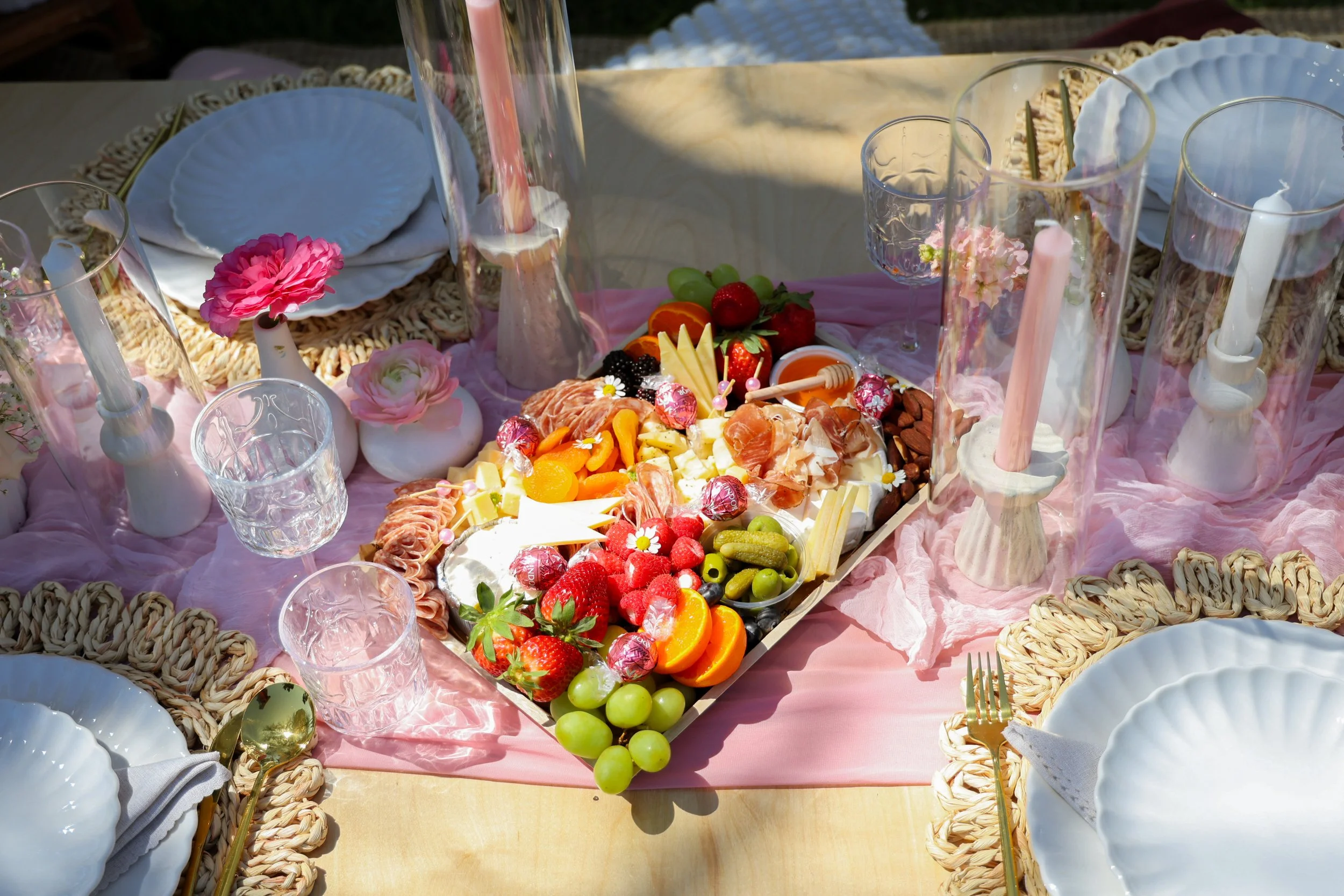 A decorative outdoor table set for a celebration with a fruit and cheese platter, pink candles in white candleholders, glassware, plates, and pink tablecloths.