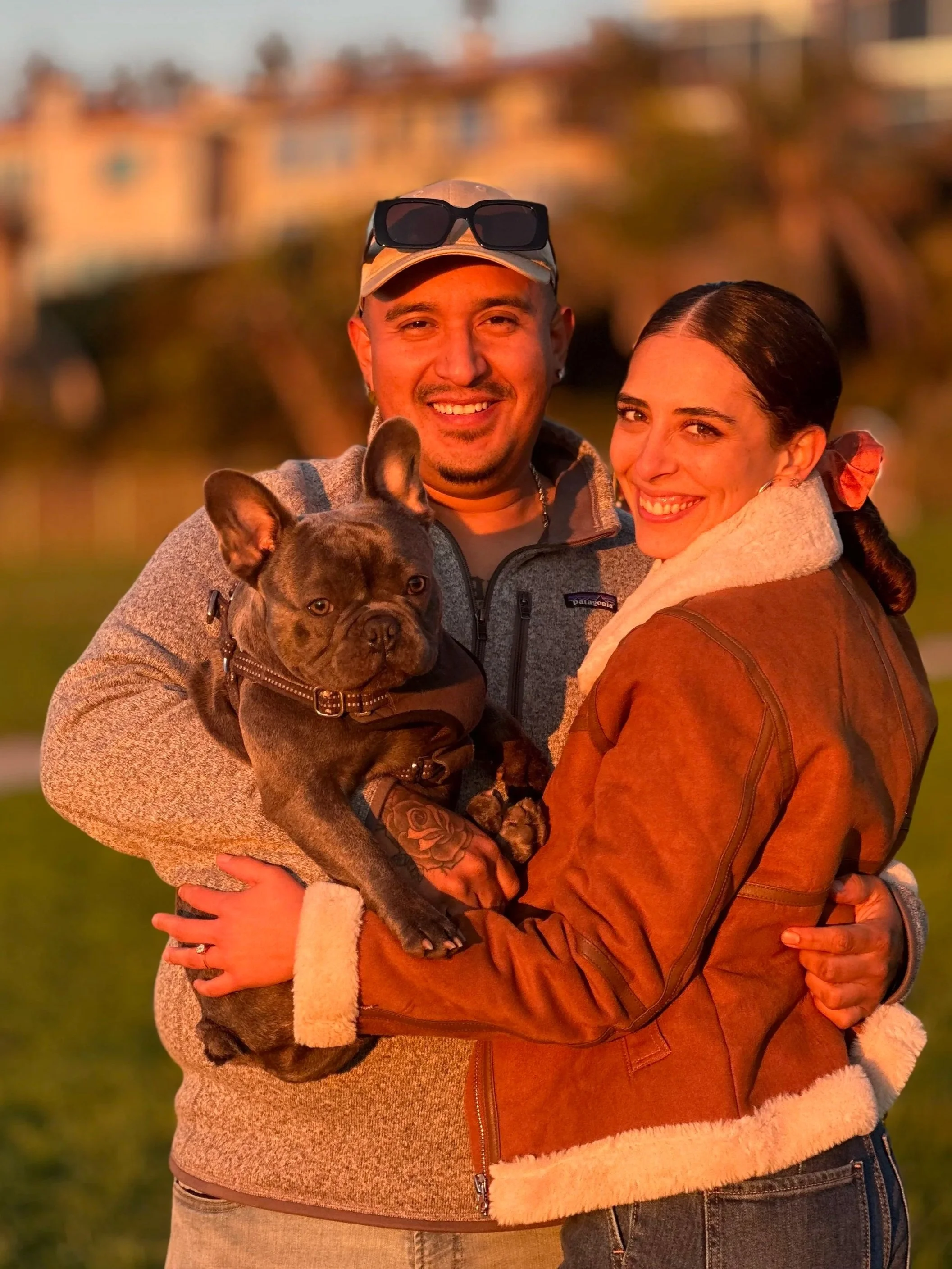 A smiling man and woman embrace outdoors during sunset, the man holding a French Bulldog puppy.