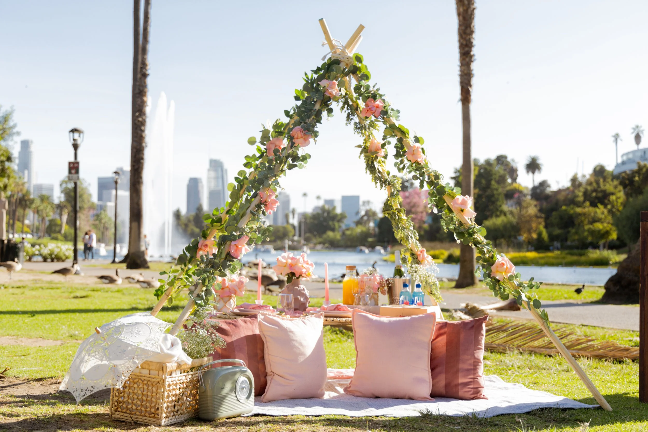 A picnic setup in a park with a floral canopy, pillows, drinks, and snacks by a water fountain and cityscape in the background.