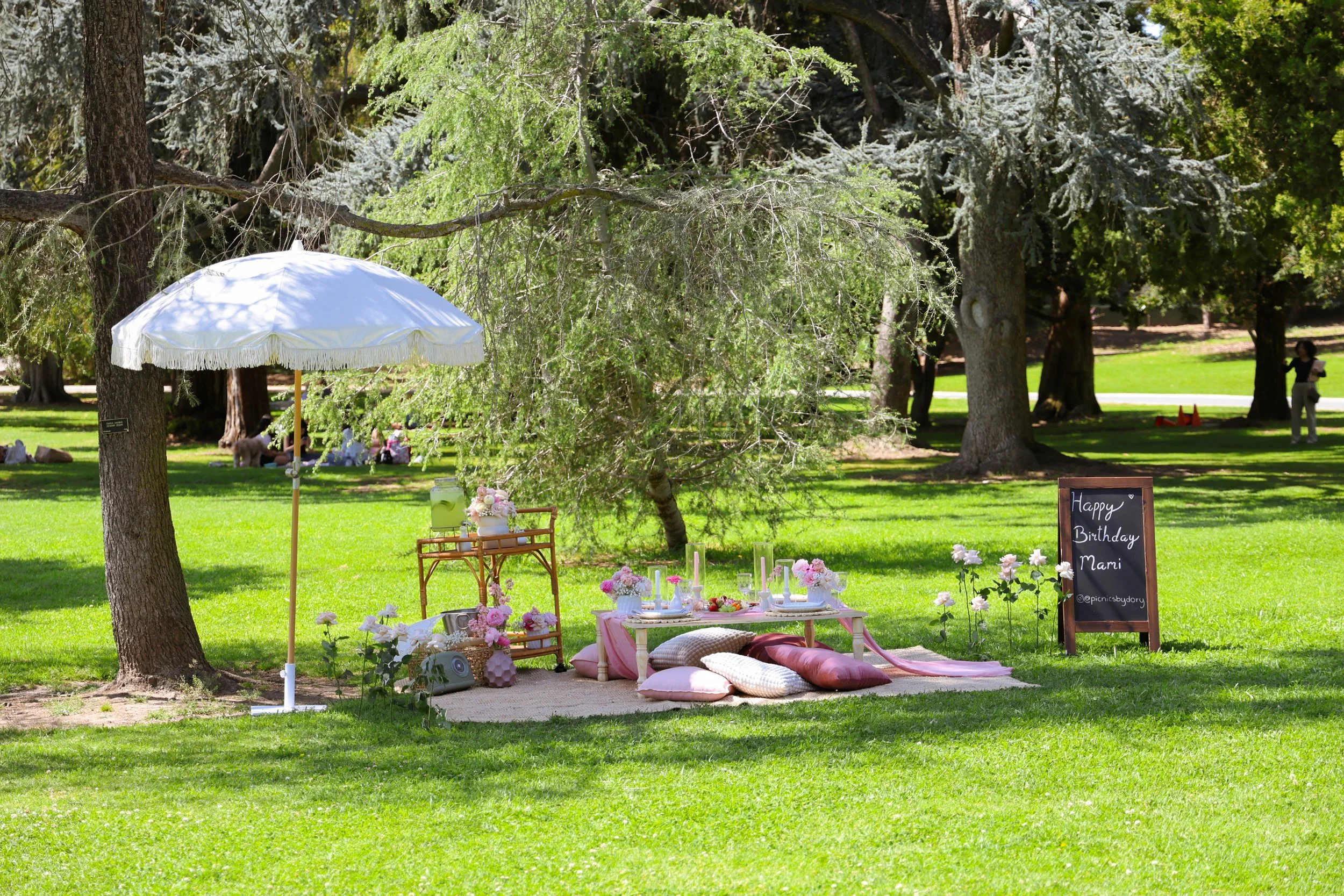 A decorated outdoor picnic setup with a pink and white theme, featuring a low table, cushions, flowers, candles, and a chalkboard sign reading "Happy Birthday Mami" in a park with trees.
