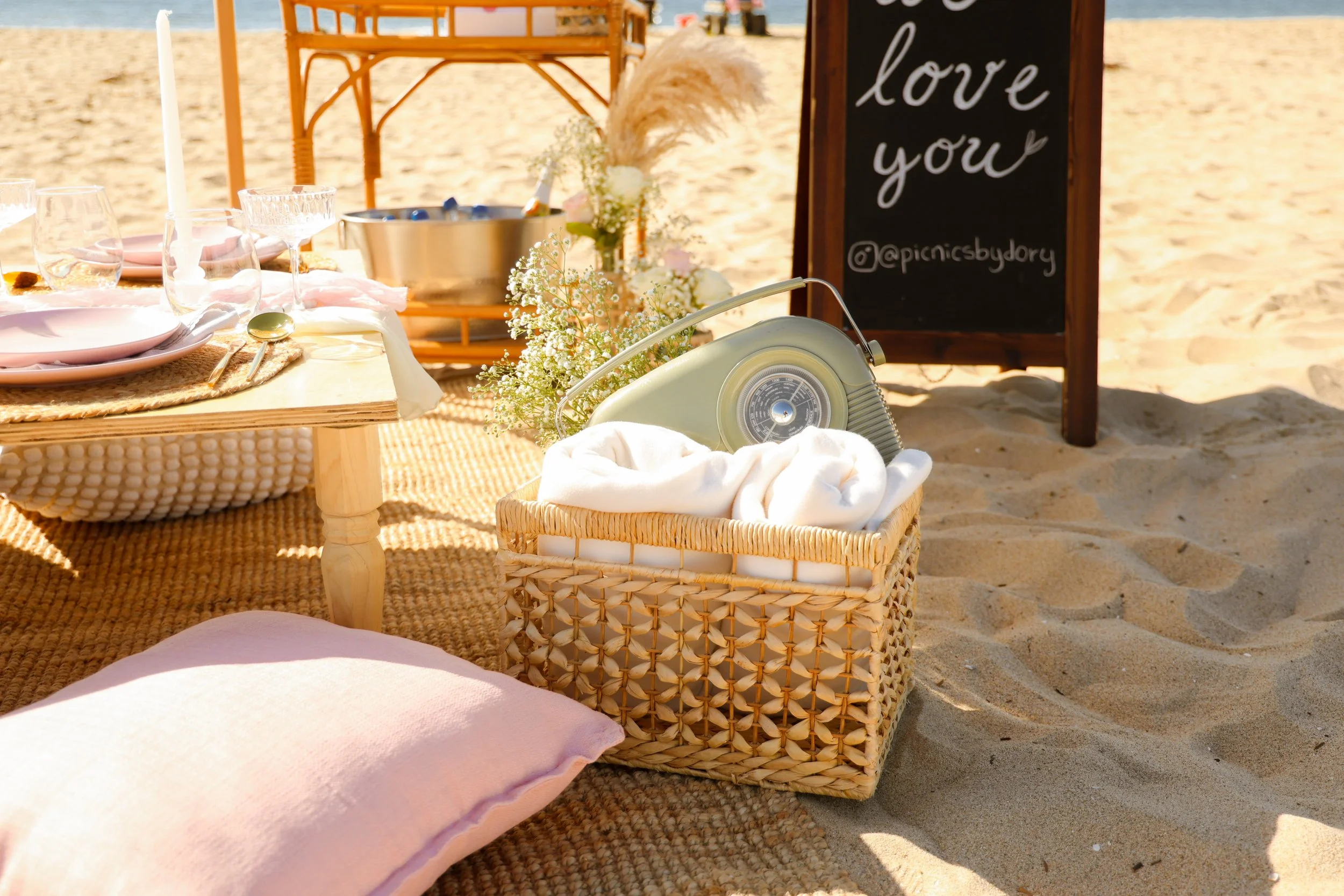 A beach picnic setup featuring a low wooden table with pink plates, glasses, and a candlestick, surrounded by woven mats and a pink pillow on sand. There is a wicker basket with a vintage radio and white blanket, floral decor, and a chalkboard sign t