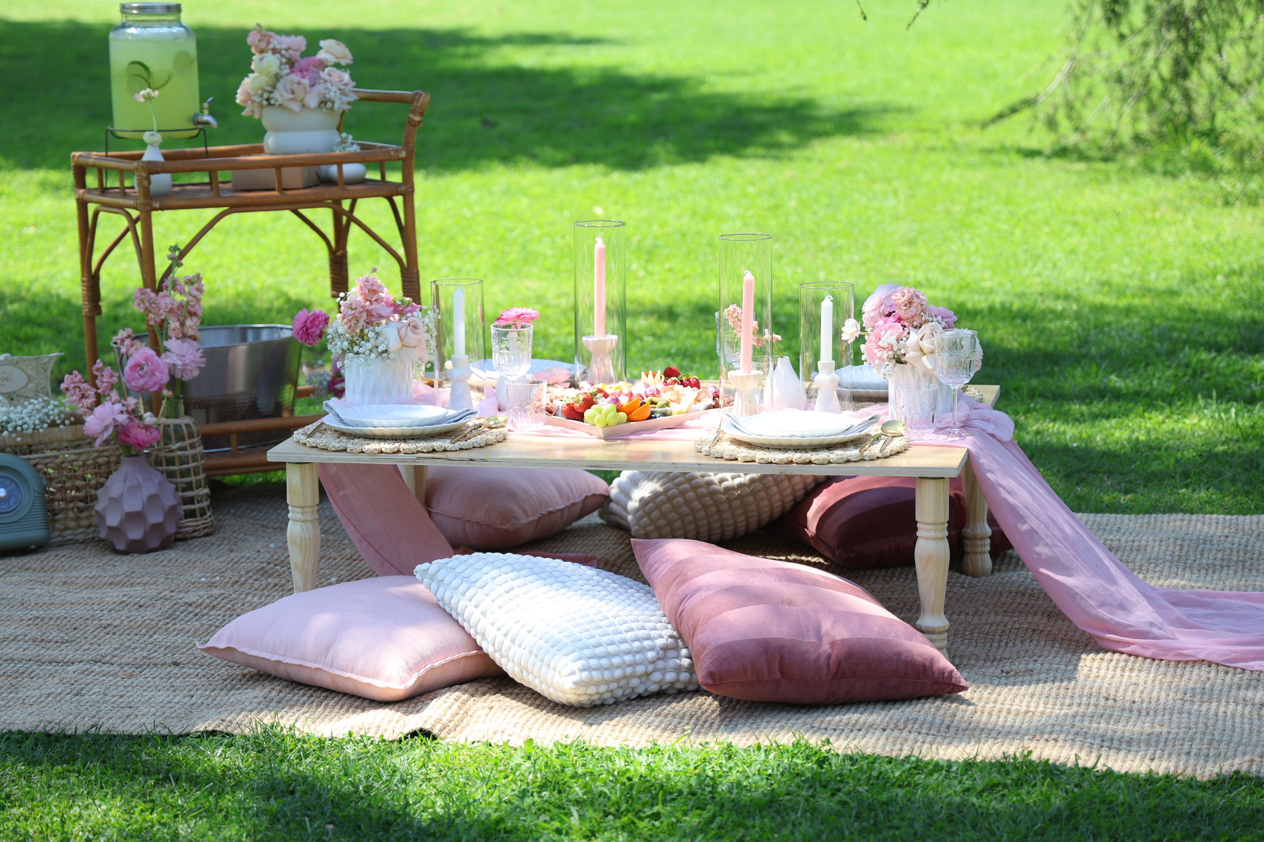 Outdoor picnic setup on a beige rug with pink and white cushions, a low wooden table set for a meal with pink candles, glasses, plates, and a plate of assorted fruit, surrounded by pink floral arrangements and a rattan cart with more flowers and a be