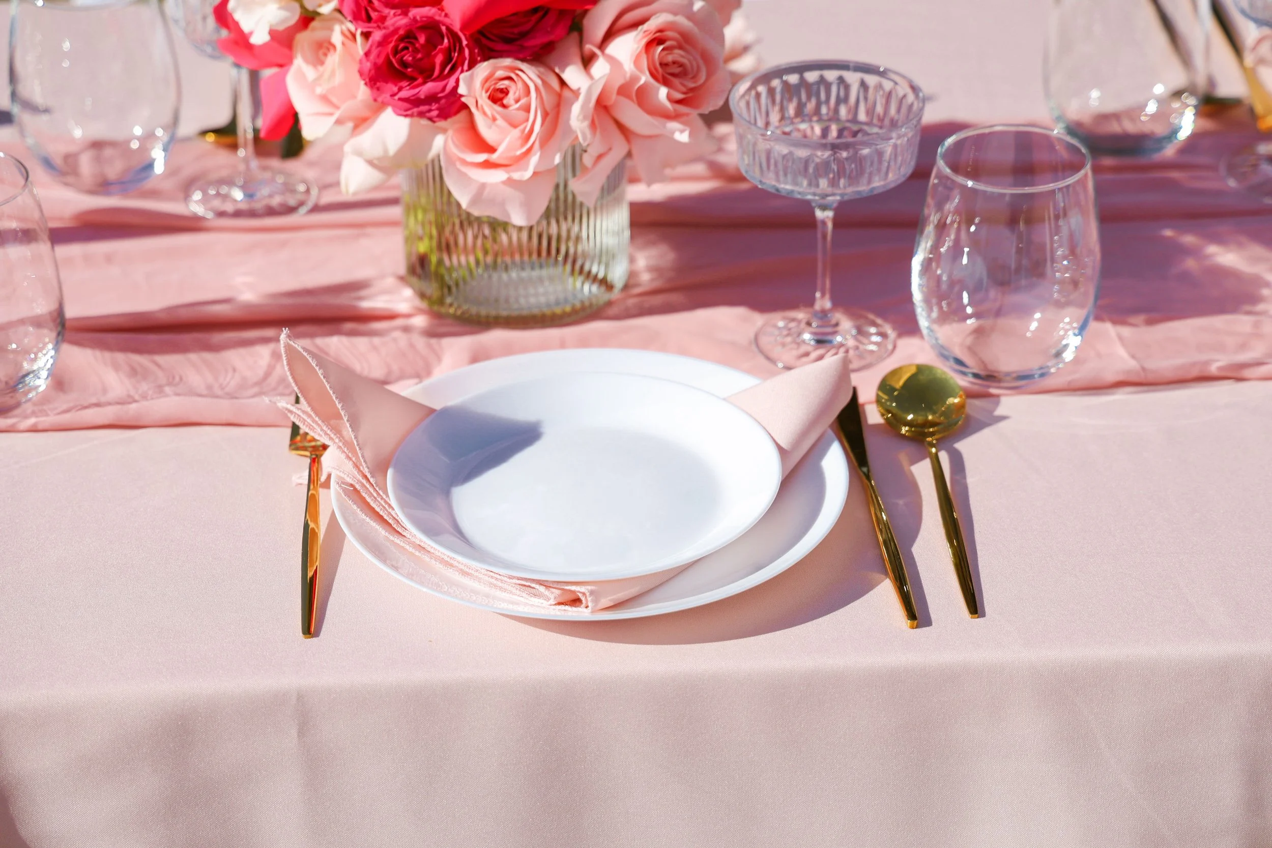 A table setting with pink tablecloth, pink napkin, white plate, gold utensils, a glass of water, a pink and gold spoon, and a flower arrangement of pink roses in a glass vase.