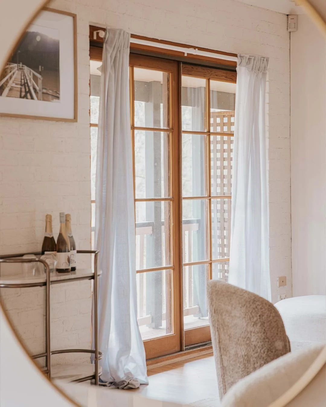A bright room with a sliding glass door leading to a balcony, white curtains, a framed photograph on the wall, a small bar cart with bottles, and upholstered chairs.