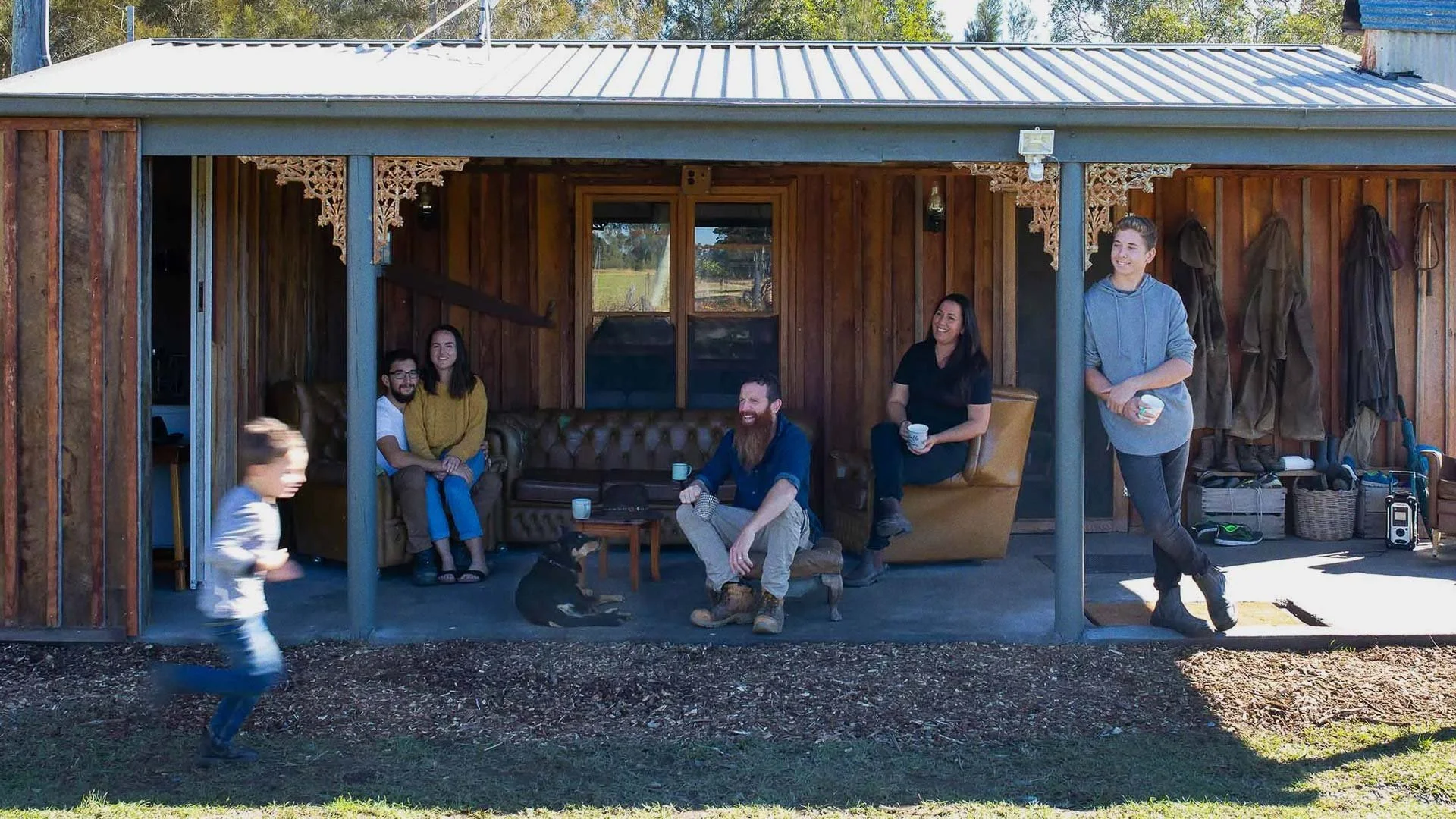 Group of five people sitting and standing on porch of rustic wooden cabin, enjoying conversation, with children and a dog present, and outdoor gear hanging on the wall.