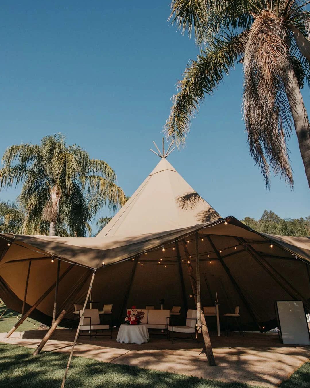 A large beige outdoor tent with string lights, surrounded by tall palm trees, under a clear blue sky.