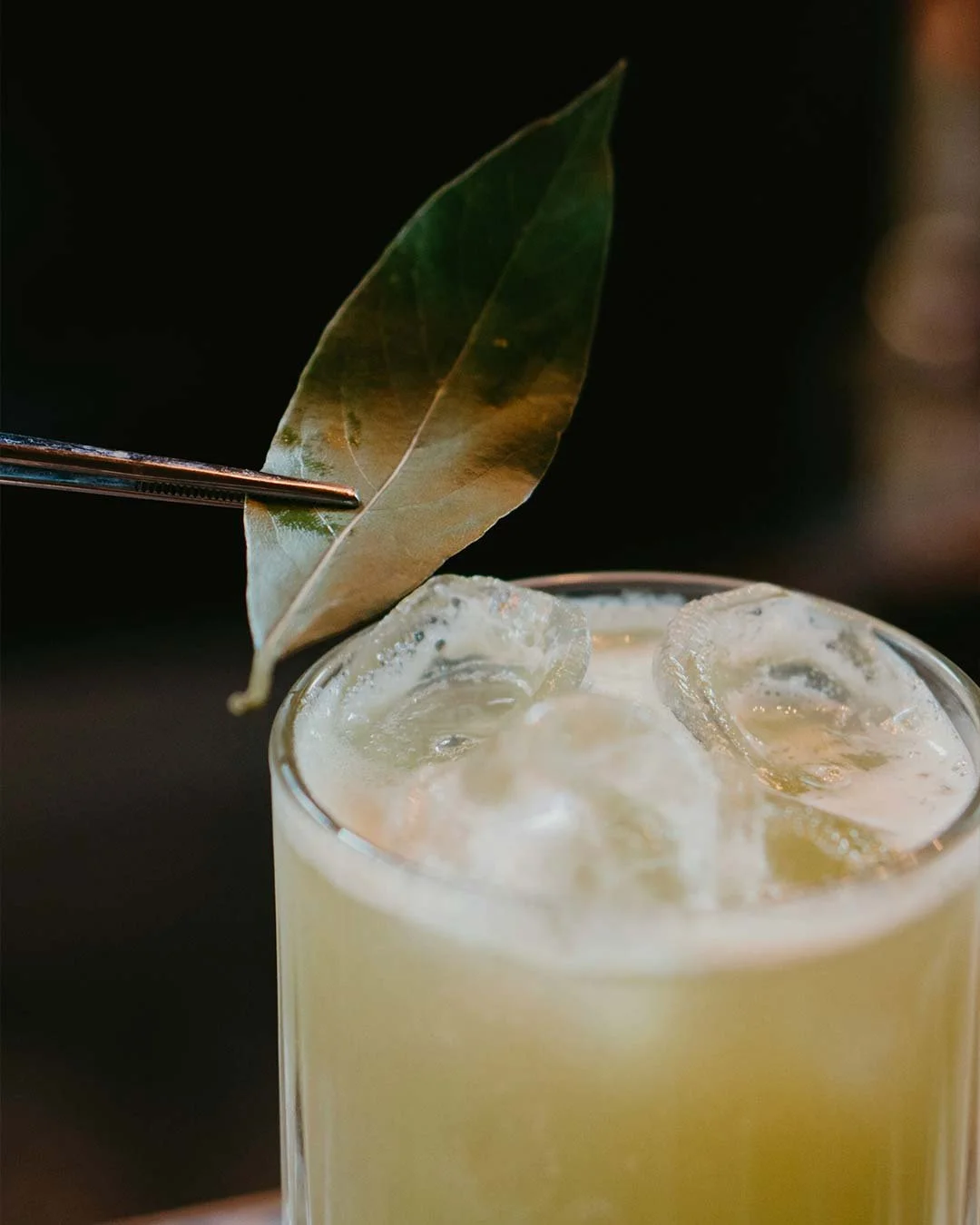 Close-up of a cocktail glass filled with a yellowish drink and ice cubes, garnished with a bay leaf held by tongs against a dark background.